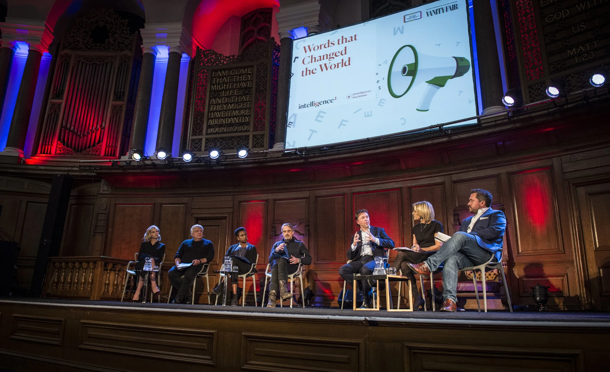 Panel discussion on stage with seven people seated in a circle, with a large screen above displaying the words "Words that Changed the World" and a megaphone graphic. The setting is a grand room with ornate architectural details and colorful lighting.