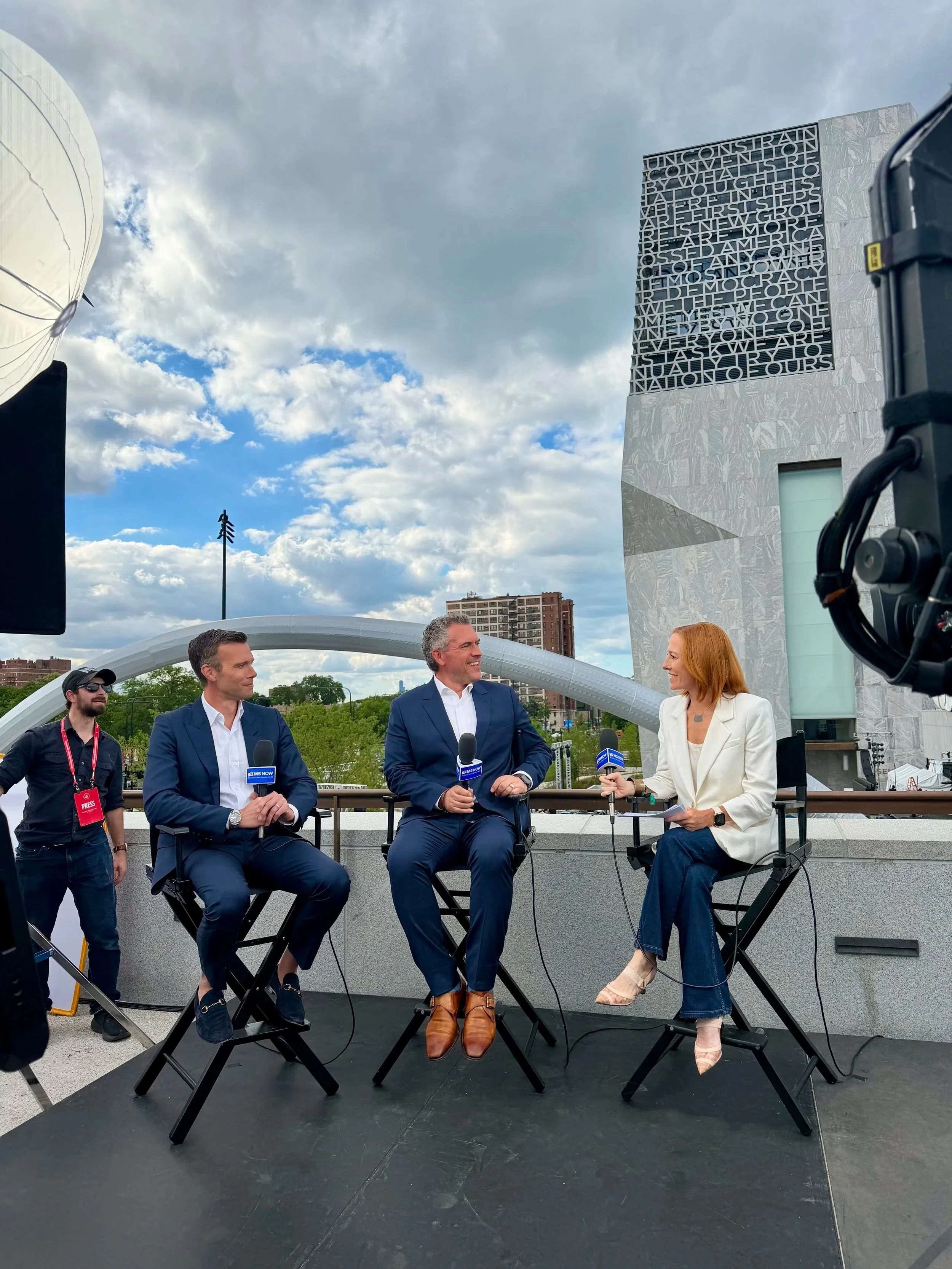 A news interview with four people sitting on a black stage outdoors, with a cityscape and a cloud-filled sky in the background. The interviewers and interviewees are holding microphones and appear to be engaged in conversation, surrounded by recording equipment and a camera.