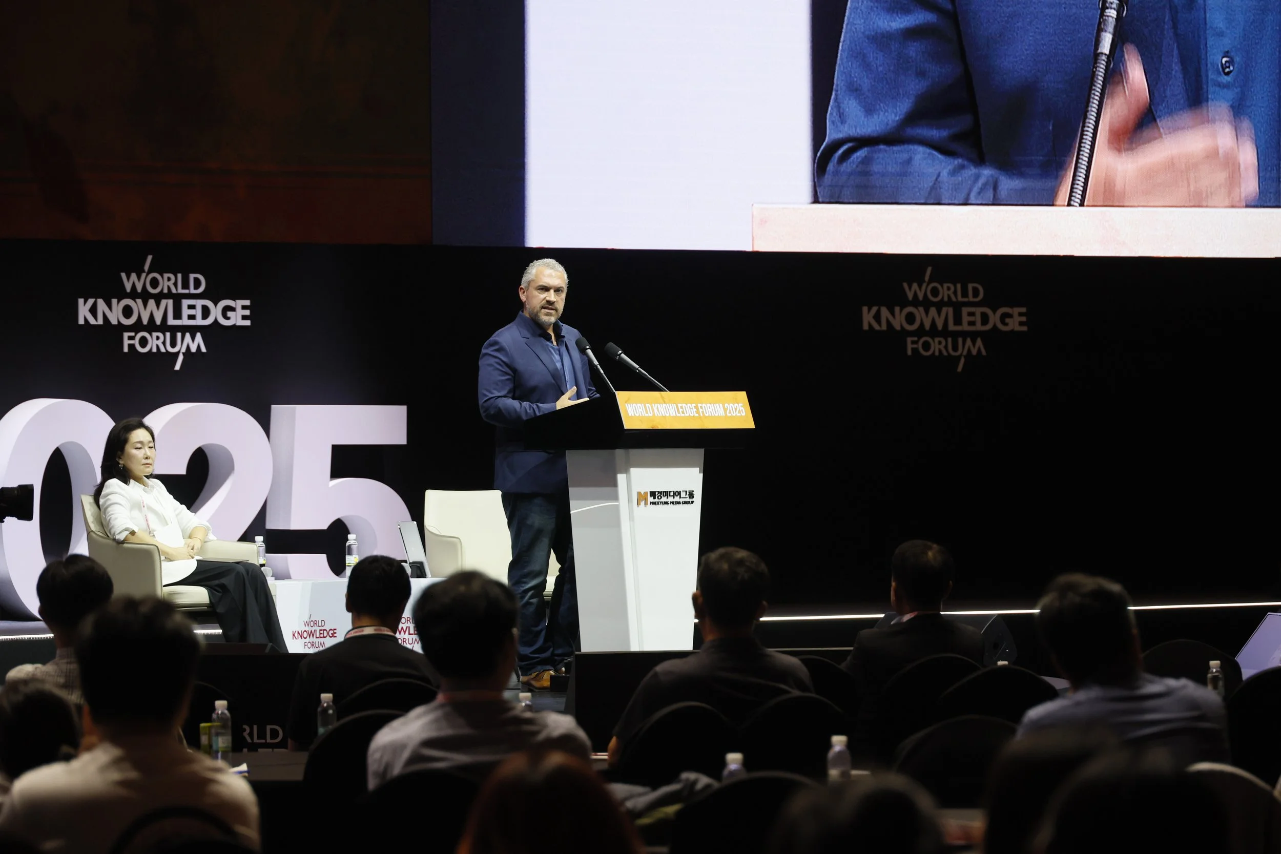A man in a blue suit is speaking at a podium during a conference at the World Knowledge Forum 2025. A woman is sitting to his left on stage, with large white letters spelling '2025' behind her. The stage background displays the event's branding and a large screen showing a close-up of the speaker.