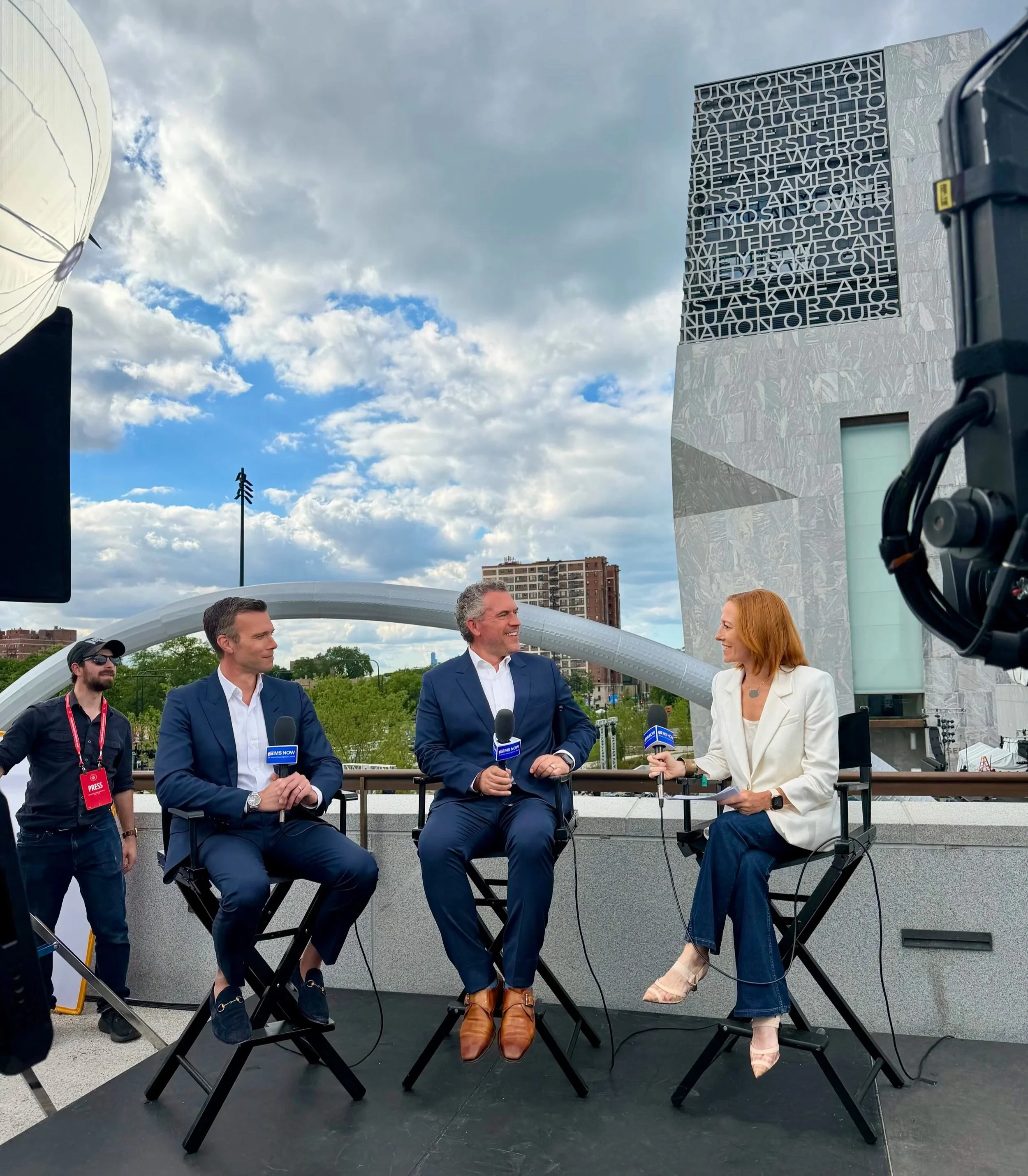 People participating in an outdoor interview or discussion, with a woman holding a microphone interviewing a man, while others watch. The scene is set against a modern building with a large digital screen displaying text, under a partly cloudy sky.