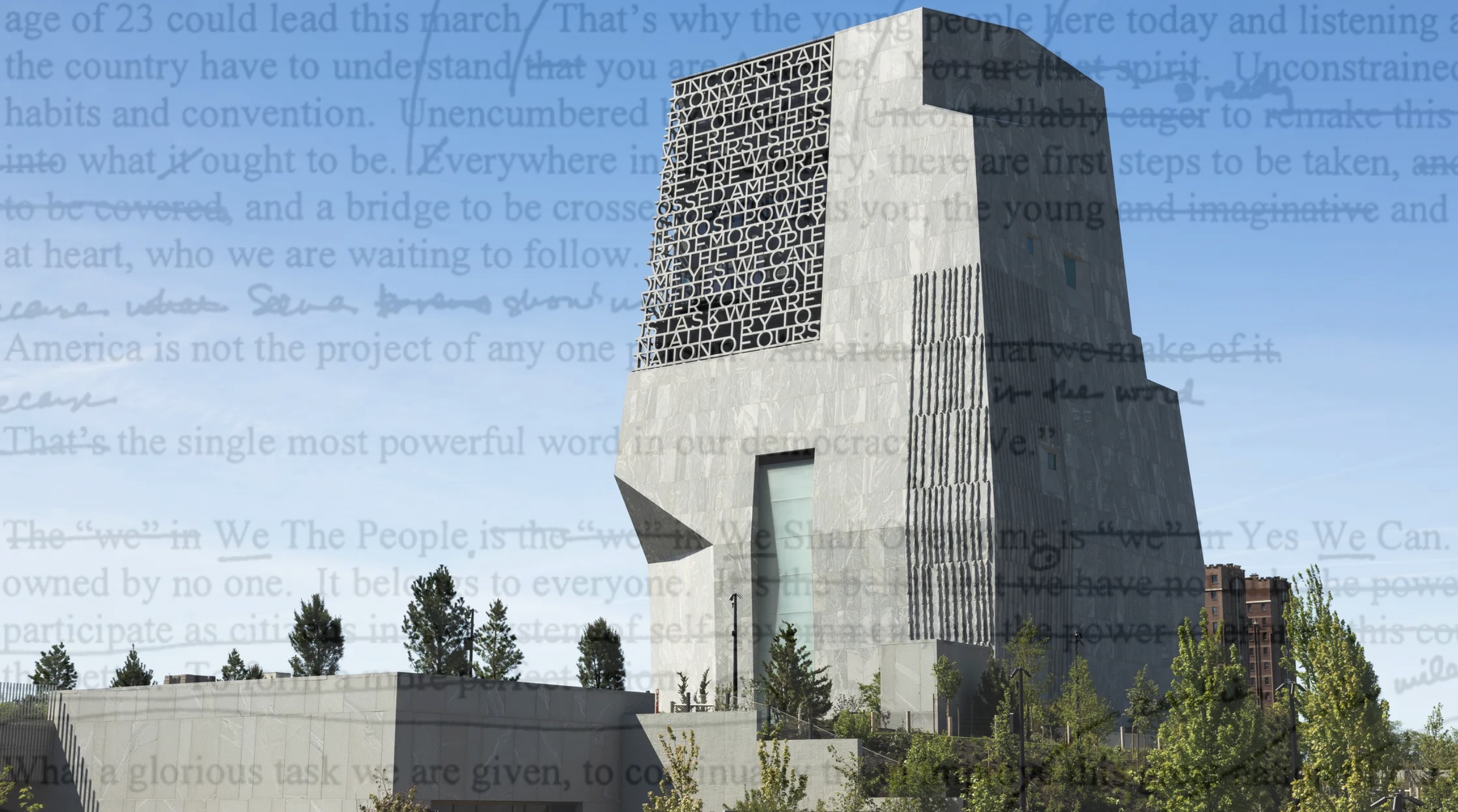 A modern, tall stone building with a black metal sculpture on the front featuring text. The sky is clear and blue, and there are small trees in front of the building.