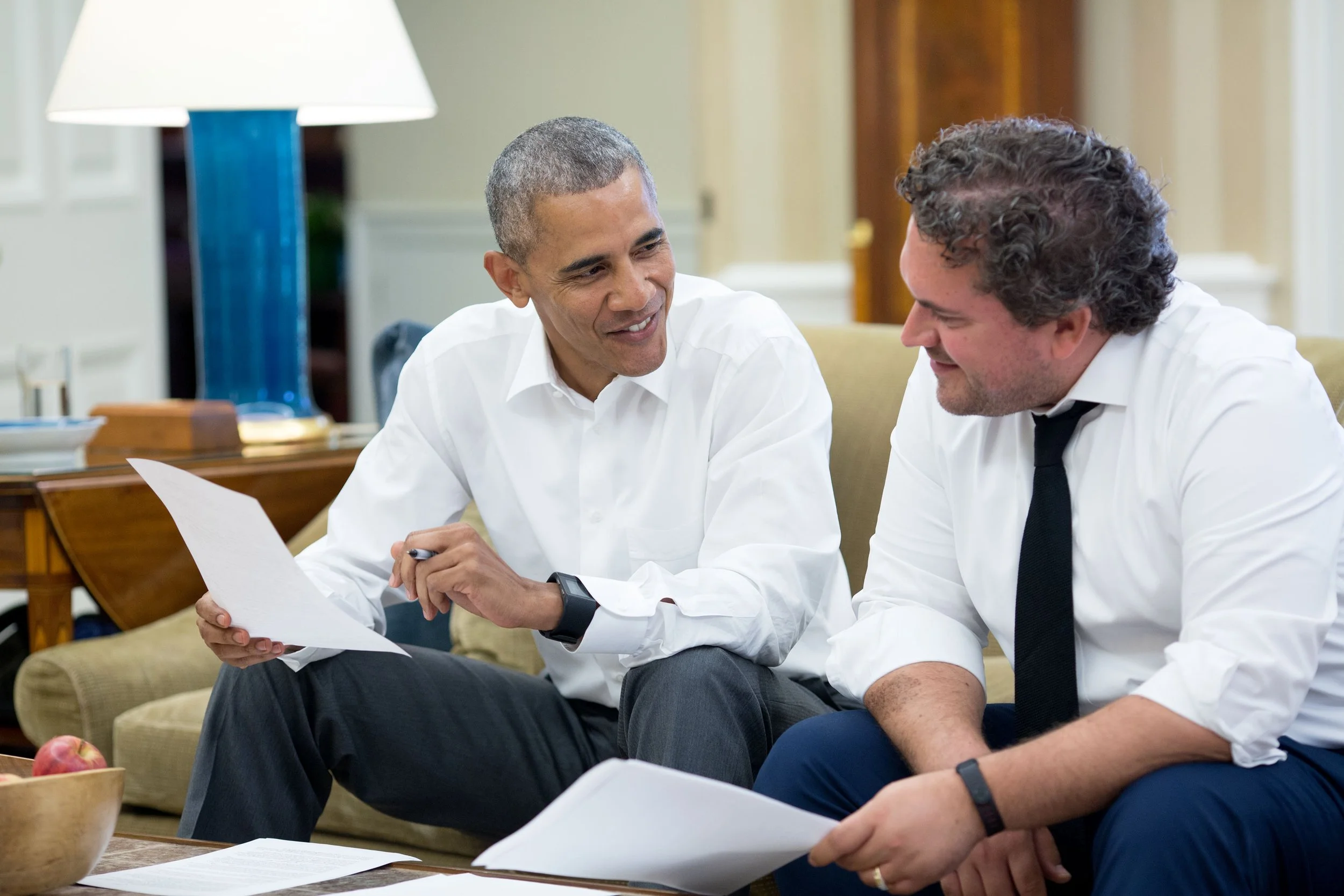Two men in business attire sitting on a beige couch, engaged in conversation and smiling, with papers in their hands, in a well-lit room with a table, a lamp, and a bowl of apples.