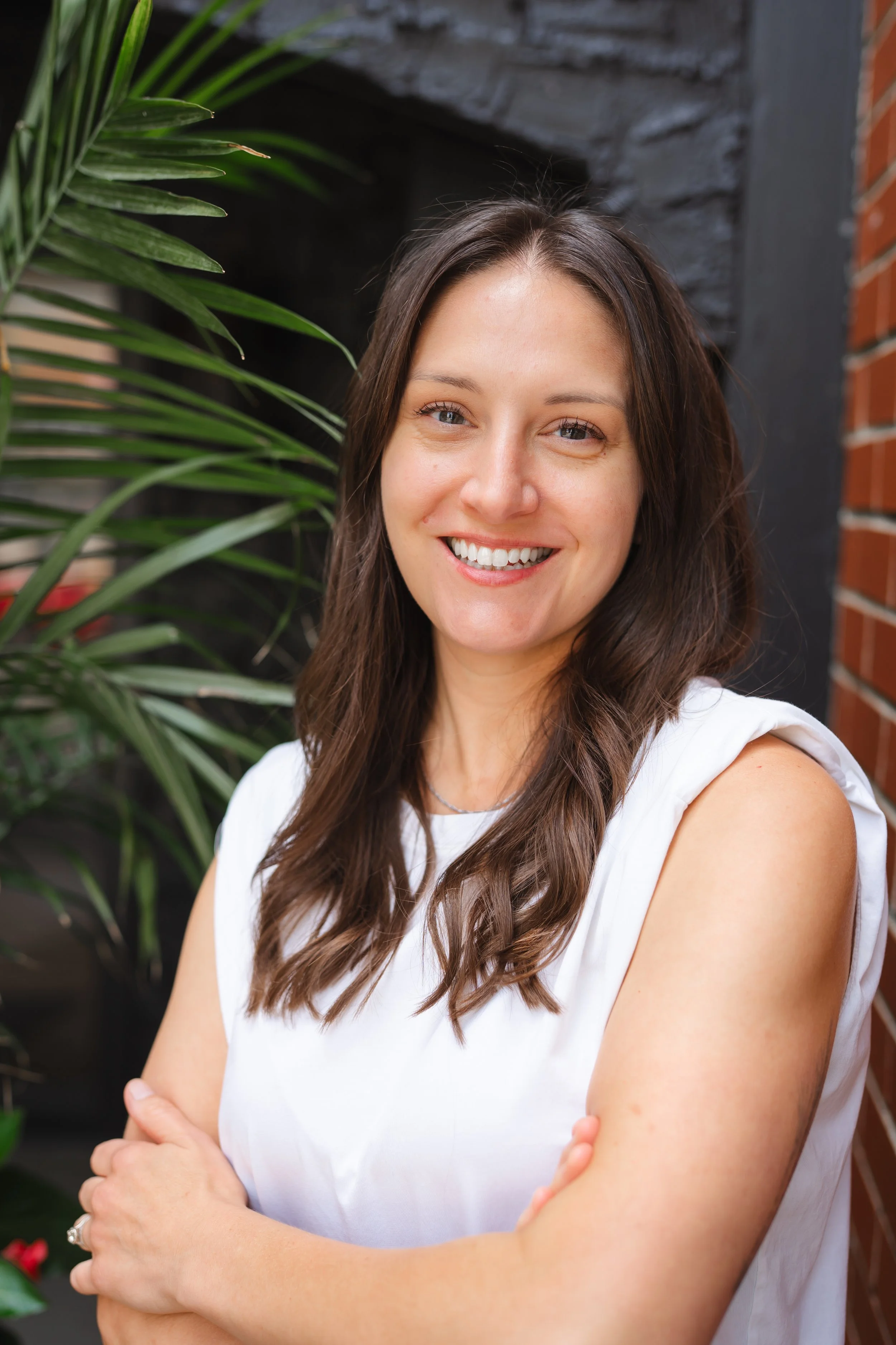 A woman with long brown hair and light skin, smiling with crossed arms, standing outdoors near green plants and a brick wall.