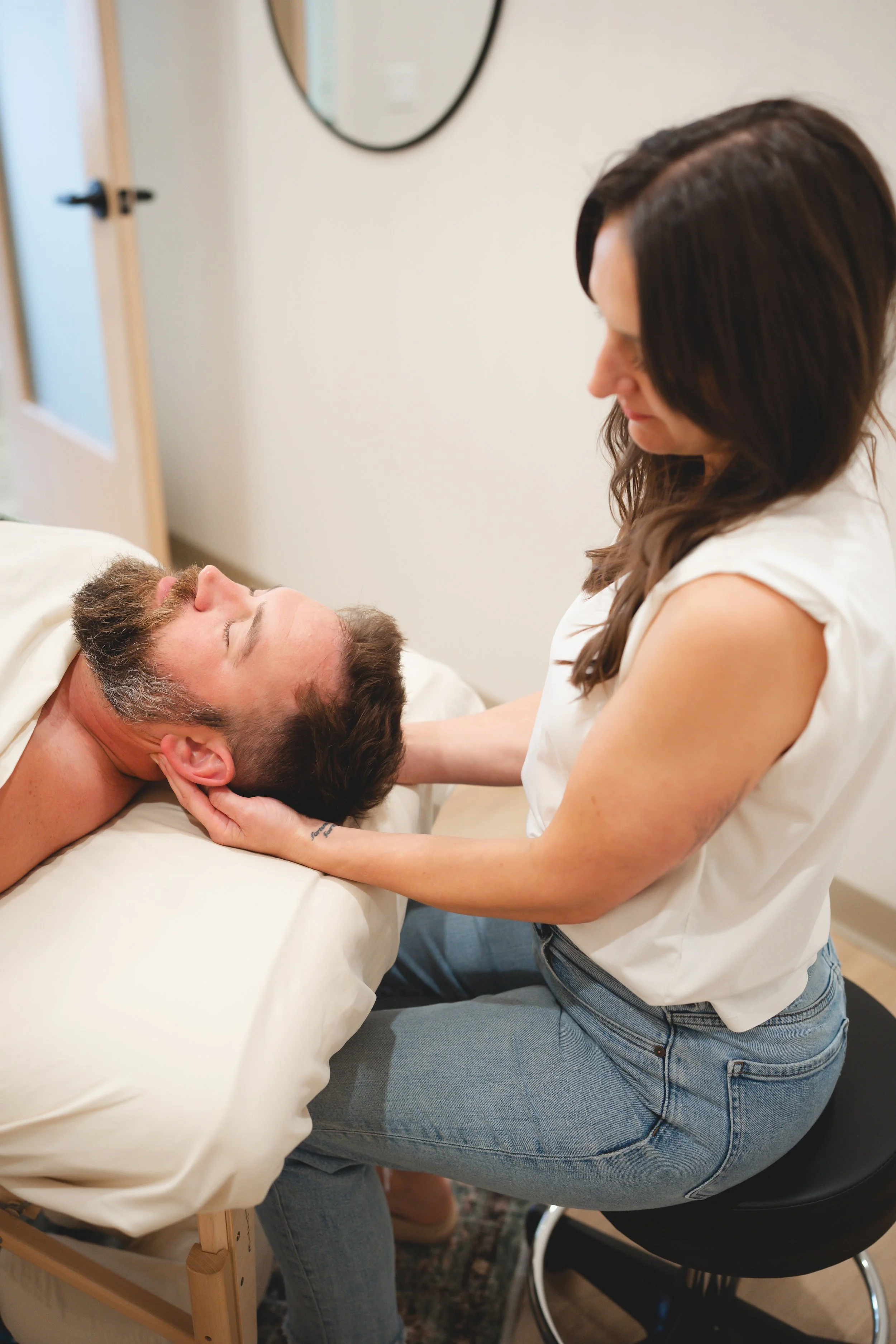 A woman providing a neck massage to a man lying on a massage table in a room with a mirror and door.