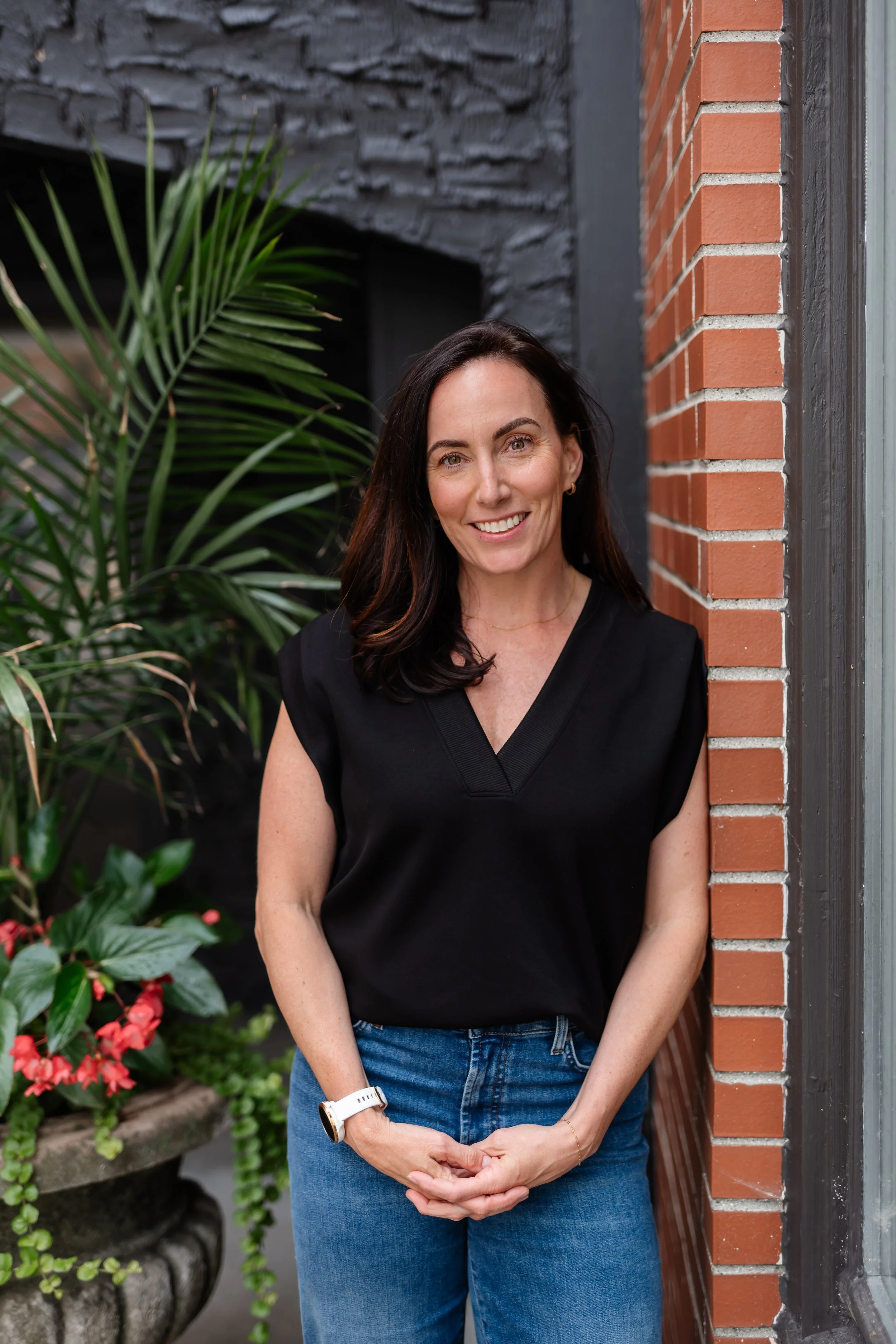 A woman with dark hair, wearing a black top and blue jeans, standing outdoors next to a brick wall and green plants, smiling at the camera.