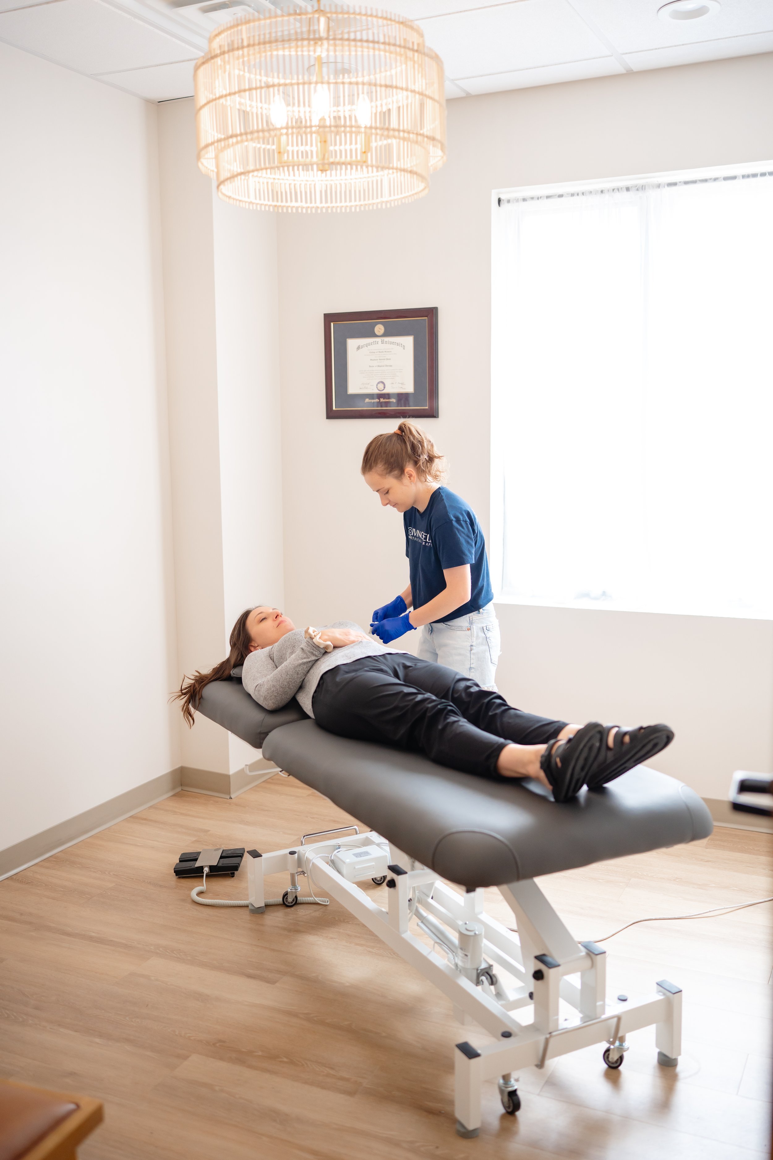 A woman lying on a medical examination table in a doctor's office, talking to a female healthcare professional wearing blue gloves and scrubs, with a diploma hanging on the wall in the background.