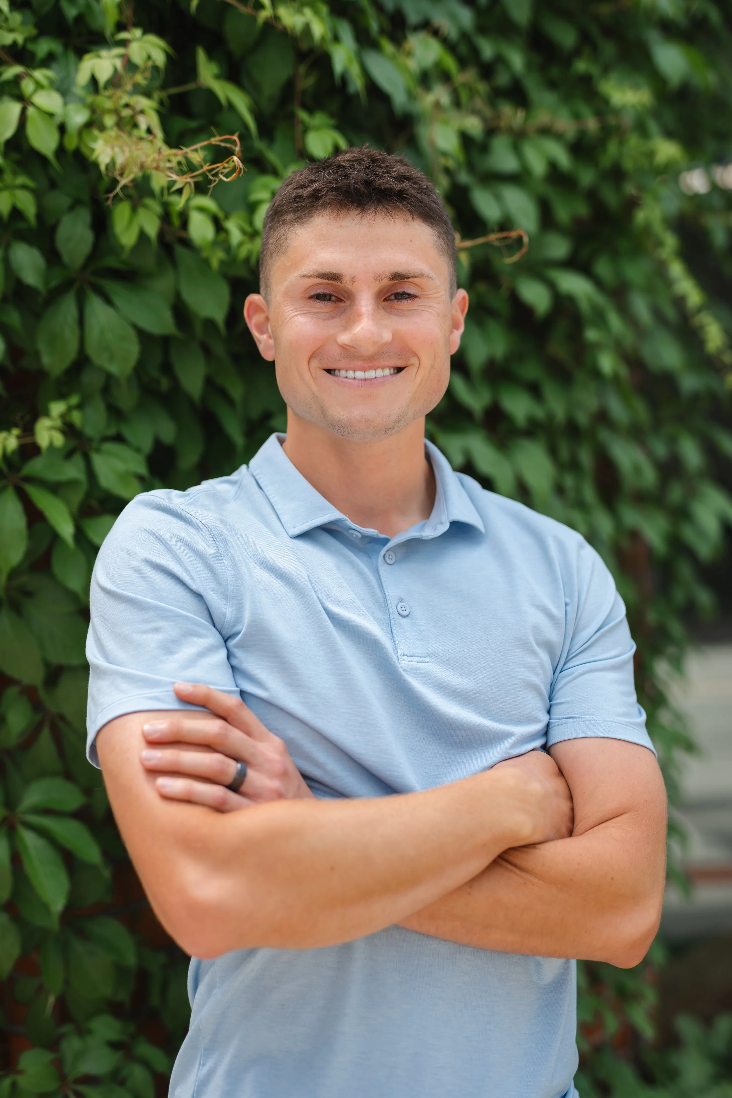 A young man in a light blue polo shirt smiling with crossed arms, standing outdoors in front of green foliage.