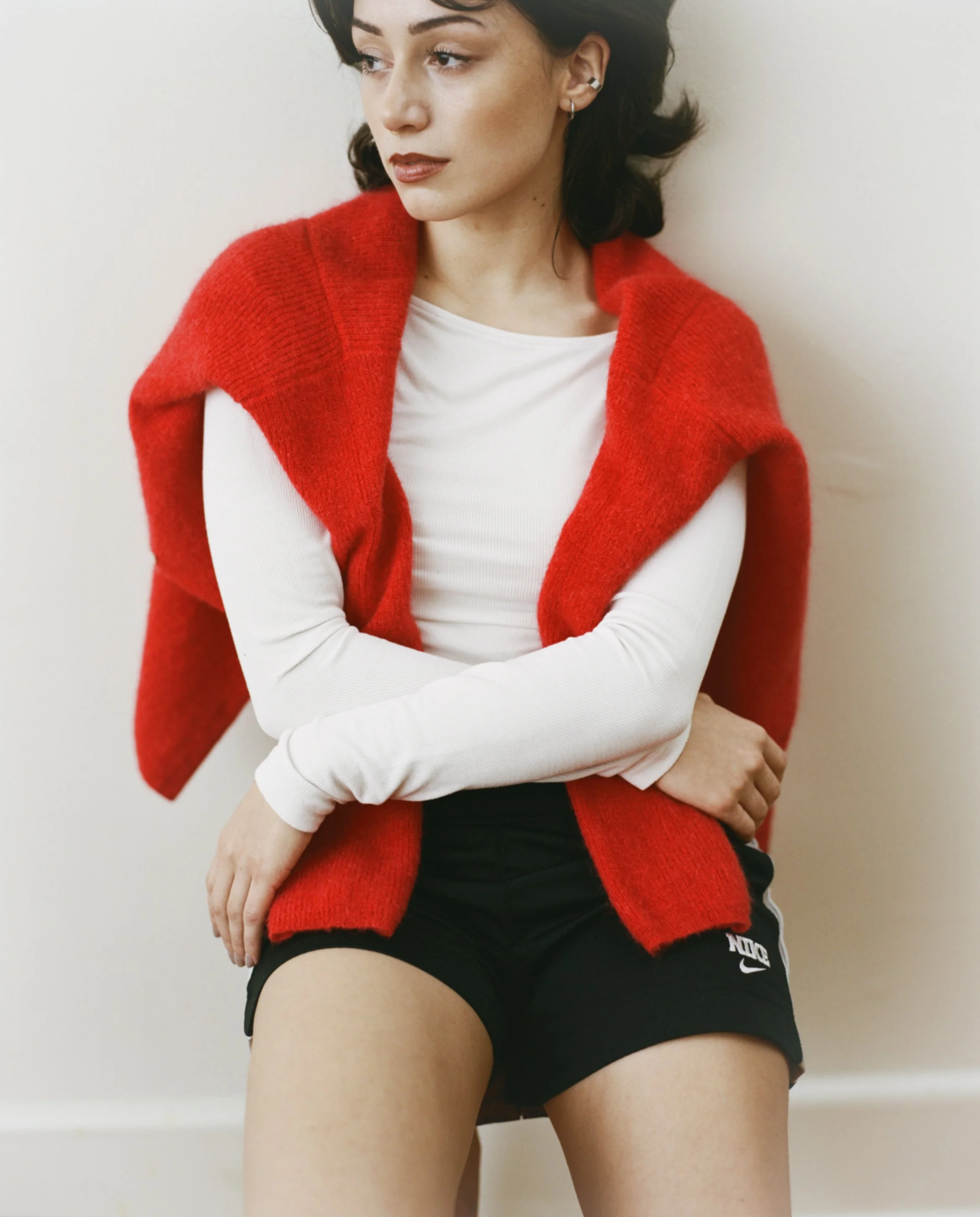 A young woman with dark hair wearing a white long sleeve top, a red sleeveless sweater draped over her shoulders, and black Nike shorts, sitting against a white wall.