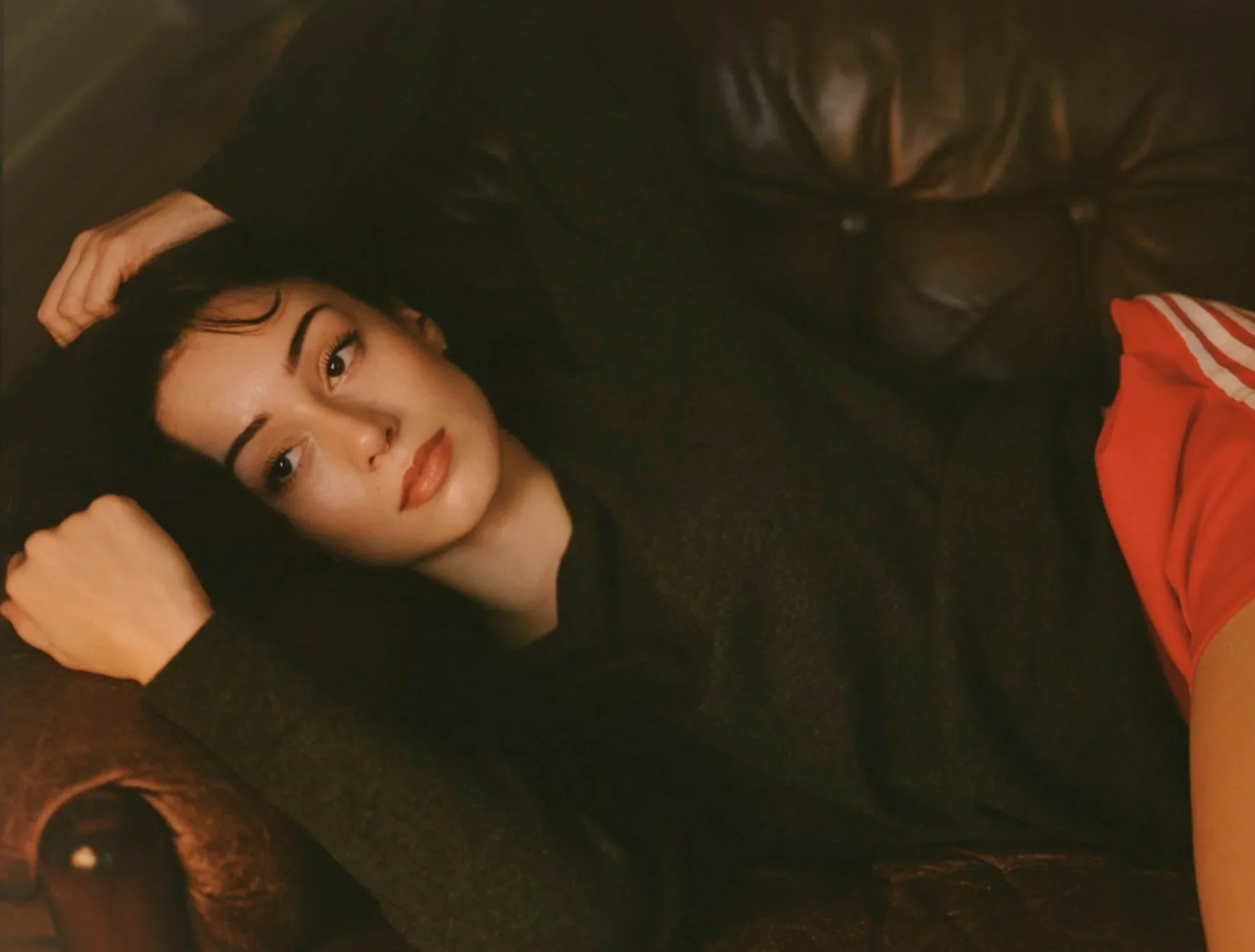 A young woman with black hair lying on a dark leather sofa, wearing a black top and red shorts with white stripes, looking directly at the camera.