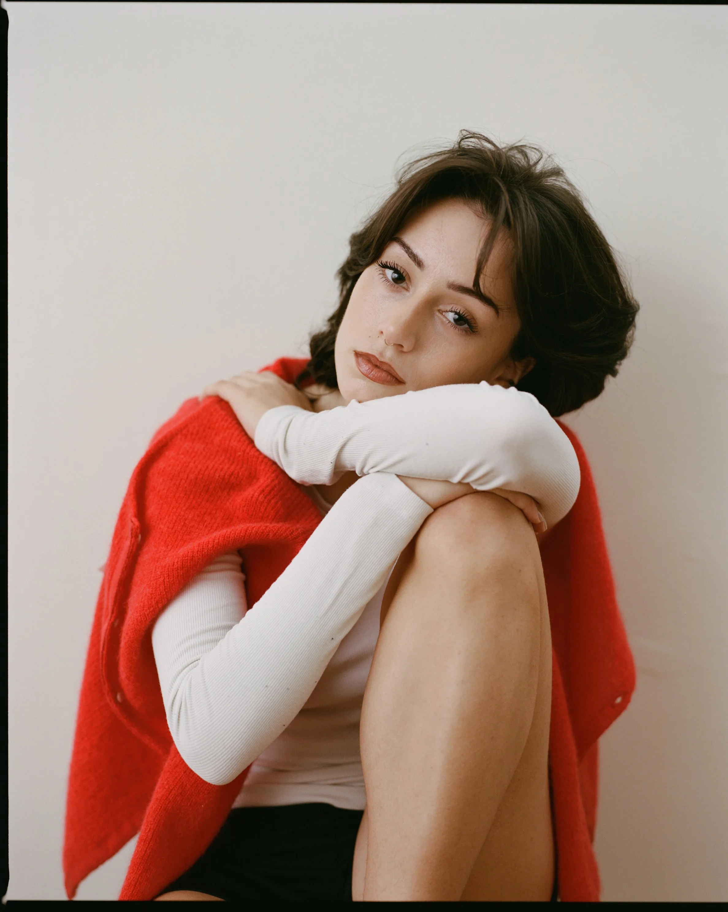 A woman with short brown hair and light makeup, wearing a red vest over a white long sleeve top, sits against a plain light-colored wall, resting her head and arms on her knee, with a contemplative expression.