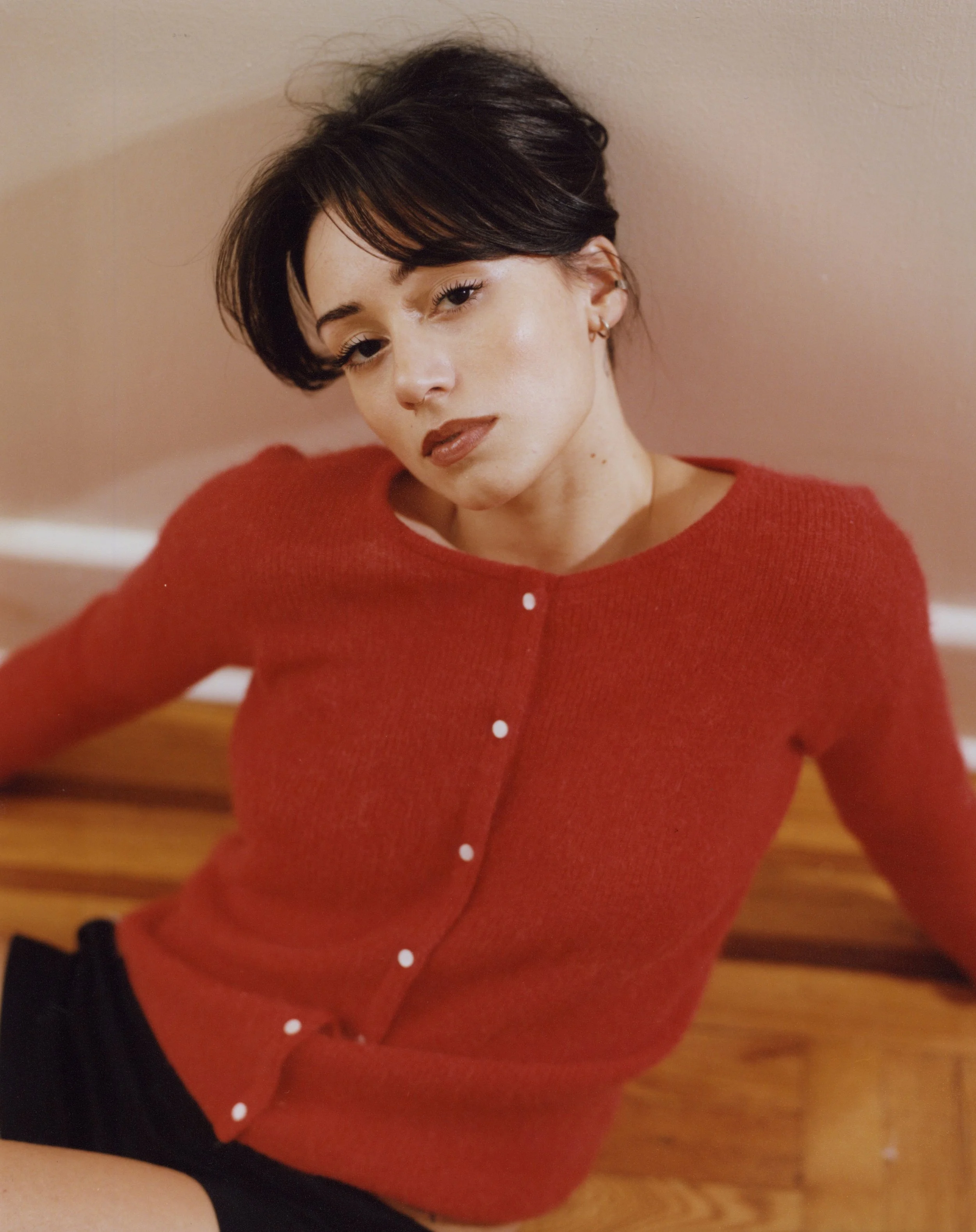 A woman with short dark hair and light skin wearing a red sweater with white buttons, sitting on a wooden surface against a pink wall.