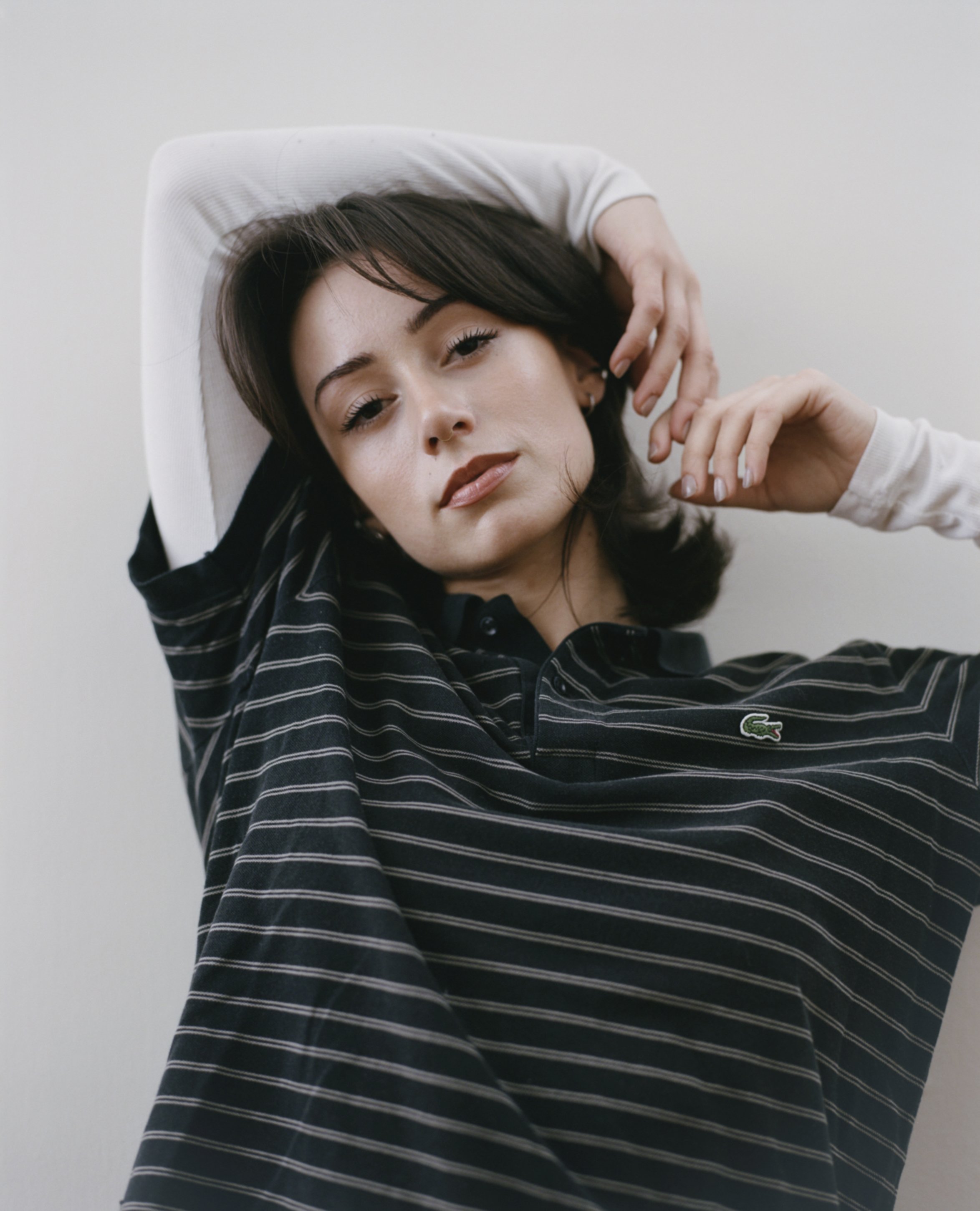 Portrait of a young woman with dark hair and light skin, wearing a black and white striped Lacoste polo shirt, posing with her hands near her face against a plain white background.