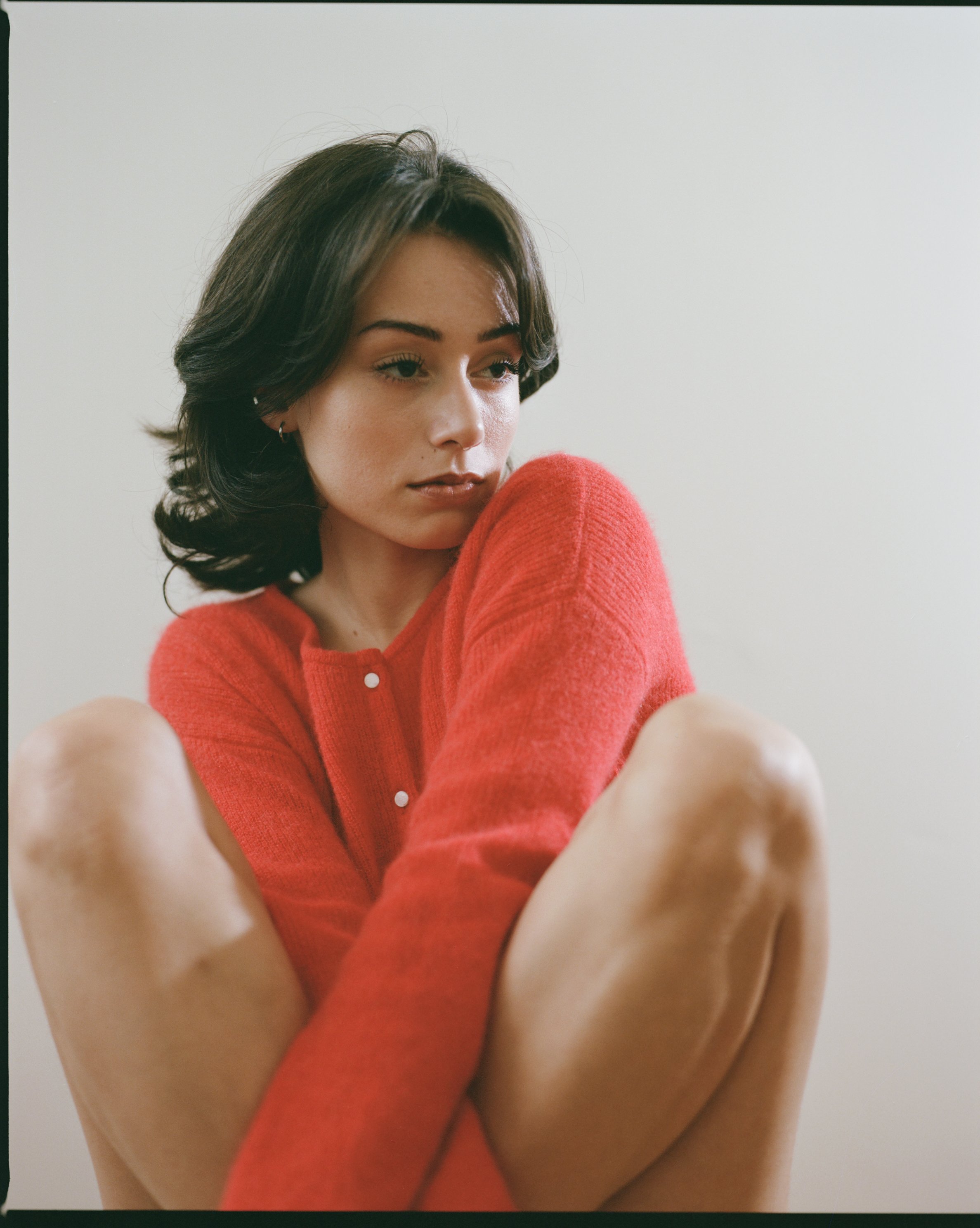 A young woman with dark hair and fair skin wearing a red sweater, sitting with her knees drawn up, against a plain white wall.