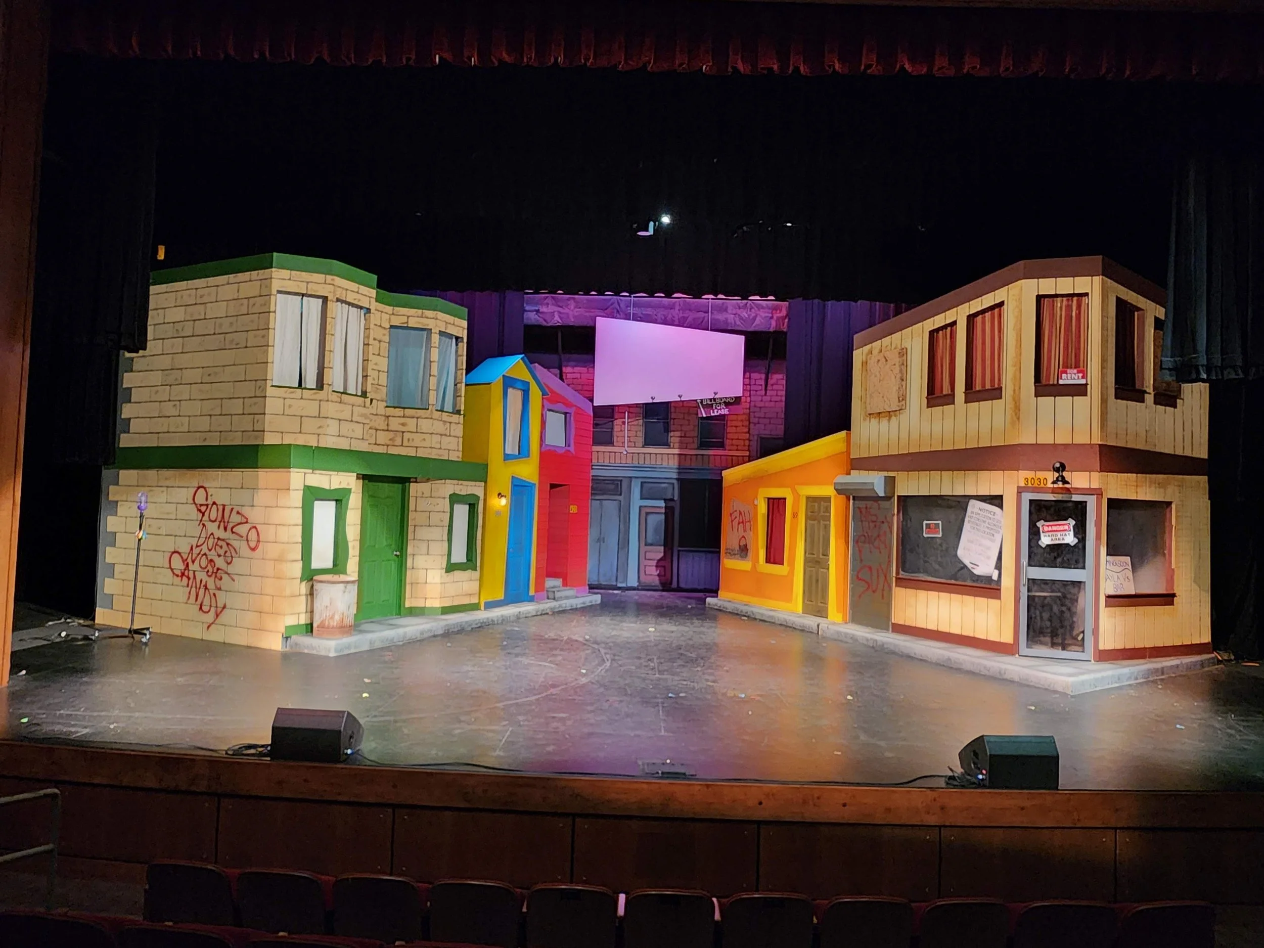 View of a stage set resembling colorful, cartoon-style buildings, with a large blank screen in the center. The stage is framed by black curtains with an ornate red valance at the top.
