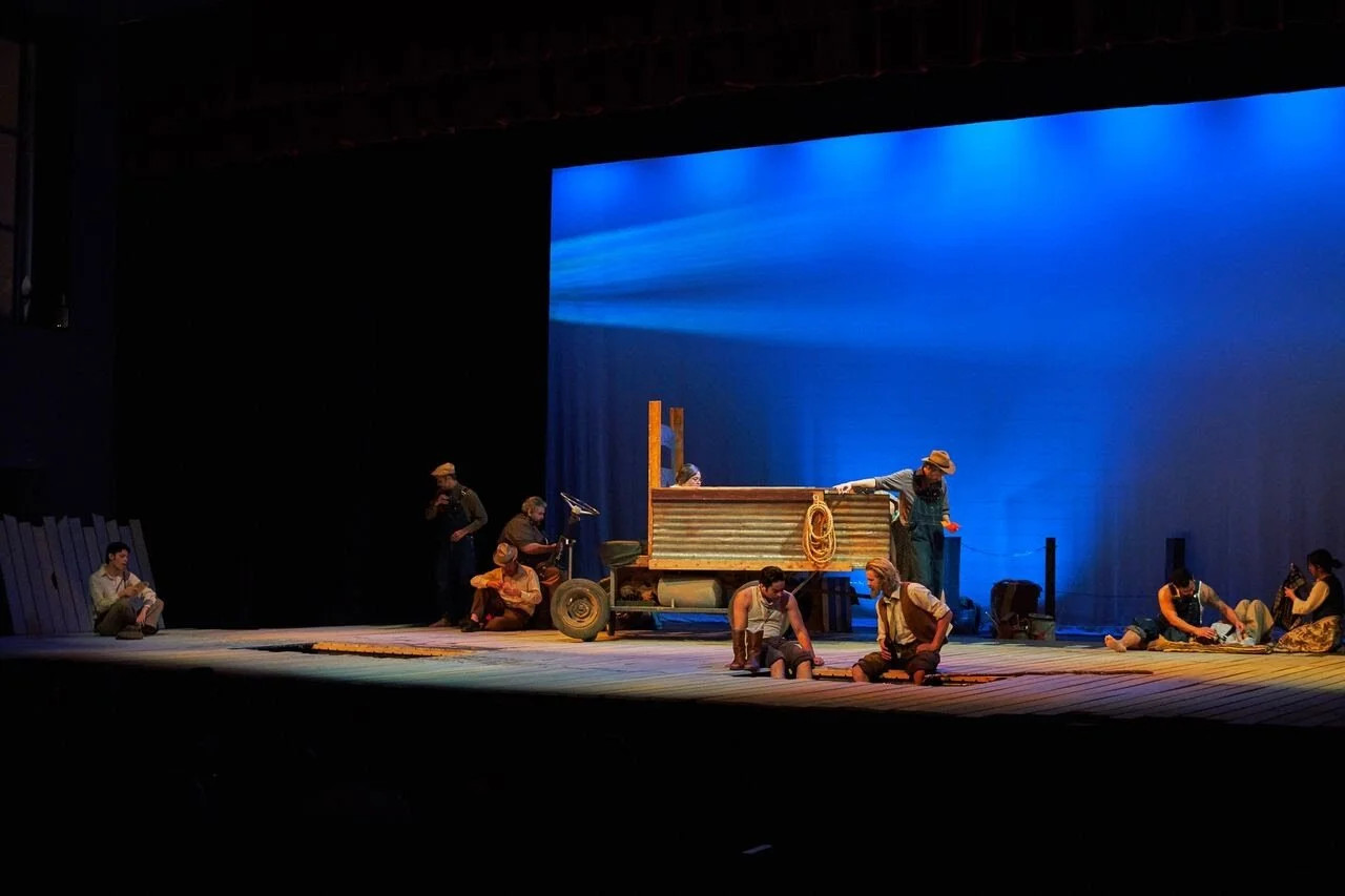 Theatrical performance of a Western scene with actors in period costumes and a small wooden wagon on stage, illuminated with blue lighting.