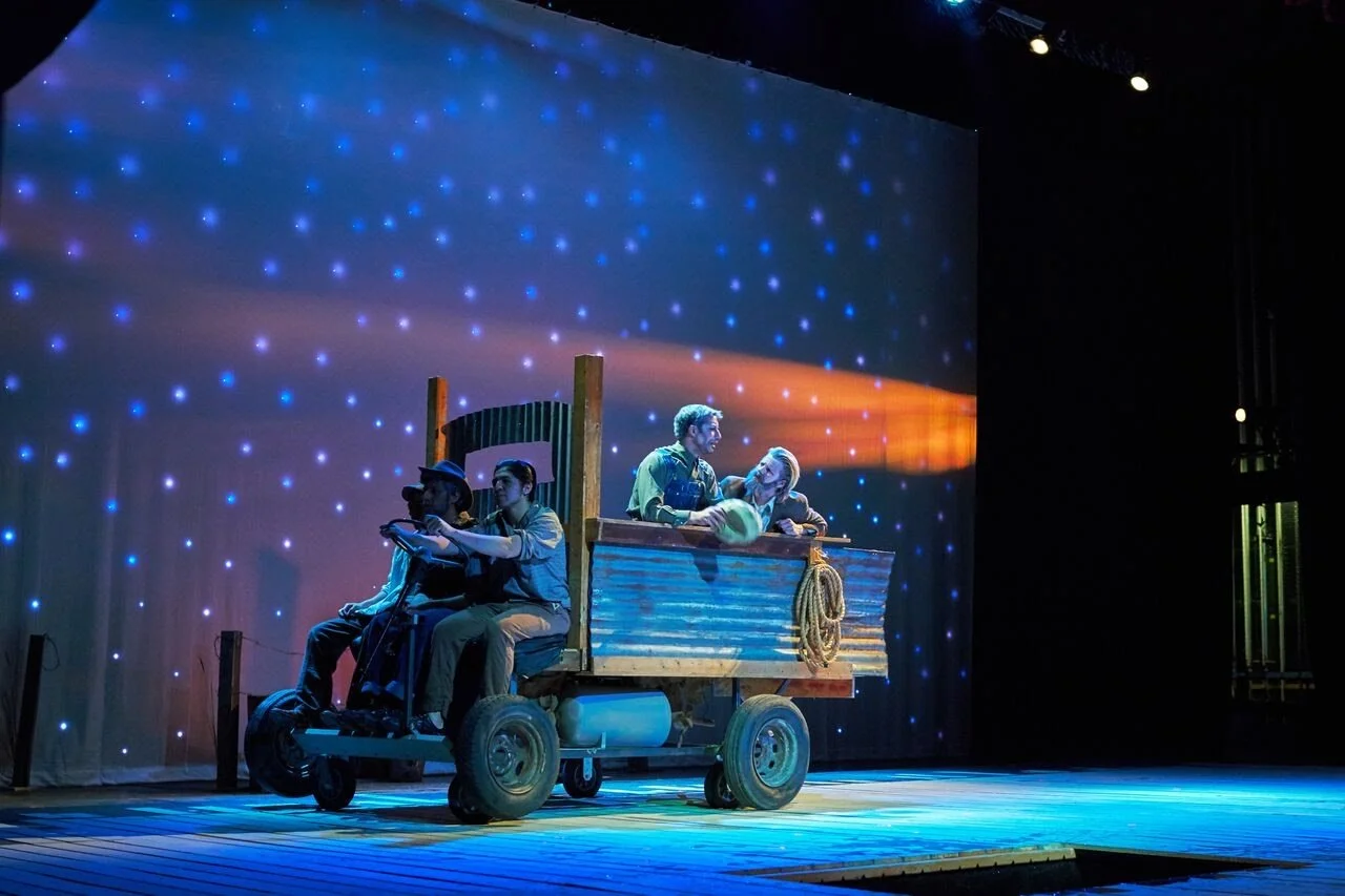 Stage scene with four actors on a wooden cart, set against a backdrop of blue starry lights, with one actor holding a steering wheel and the others engaged in conversation.