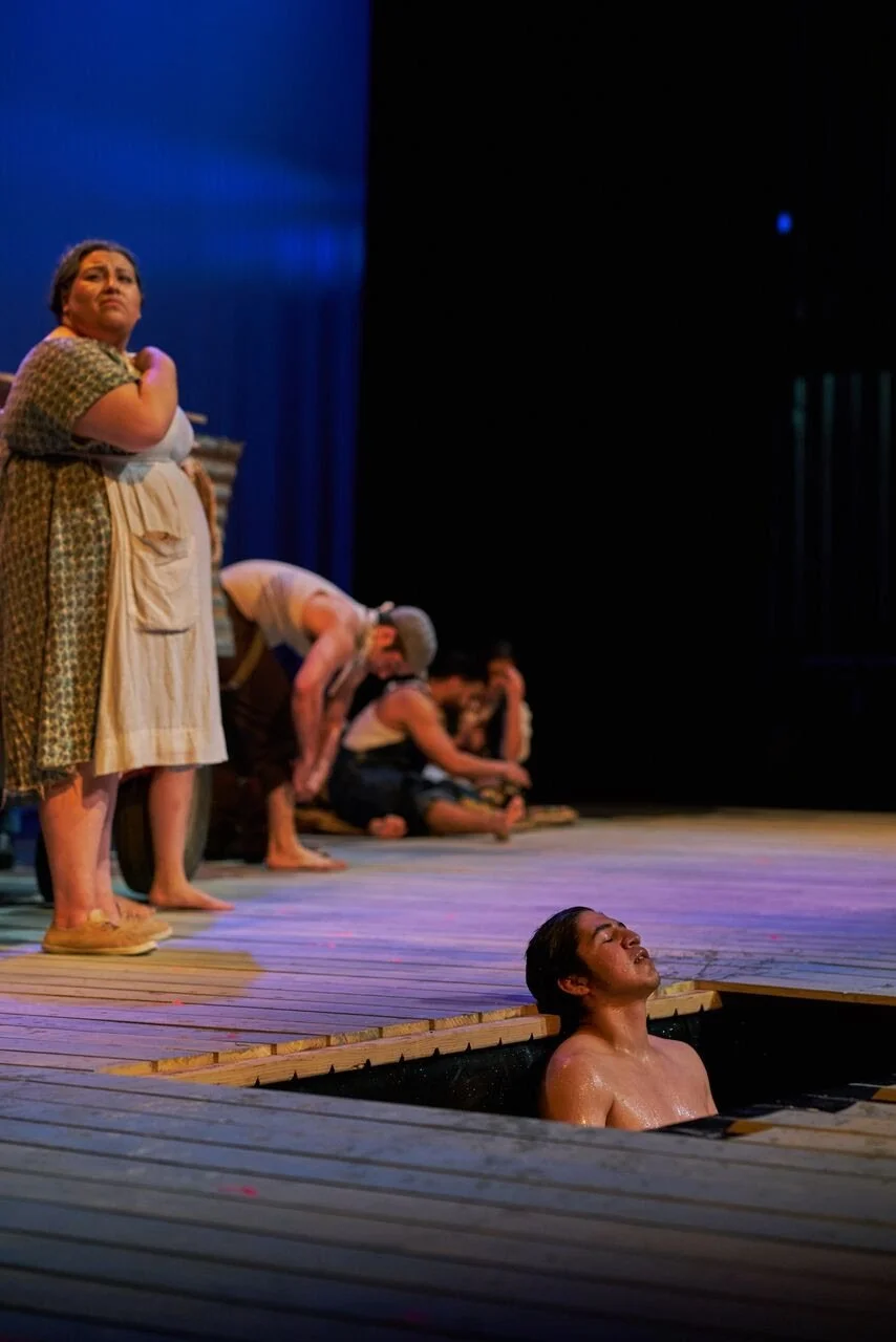 A scene from a stage play featuring a woman standing on stage, a person sitting on the floor, and a man in a small boat on the stage, with other performers in the background.