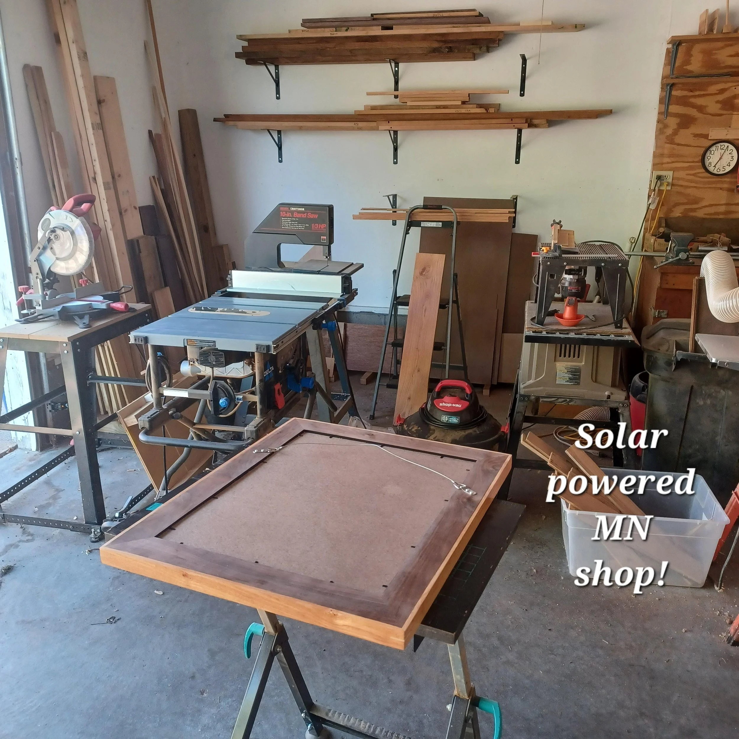 Woodworking workshop with a product in process, a table saw, and woodworking tools. Shelves with wood inventory mounted on the wall. The text overlay reads 'Solar powered shop!'
