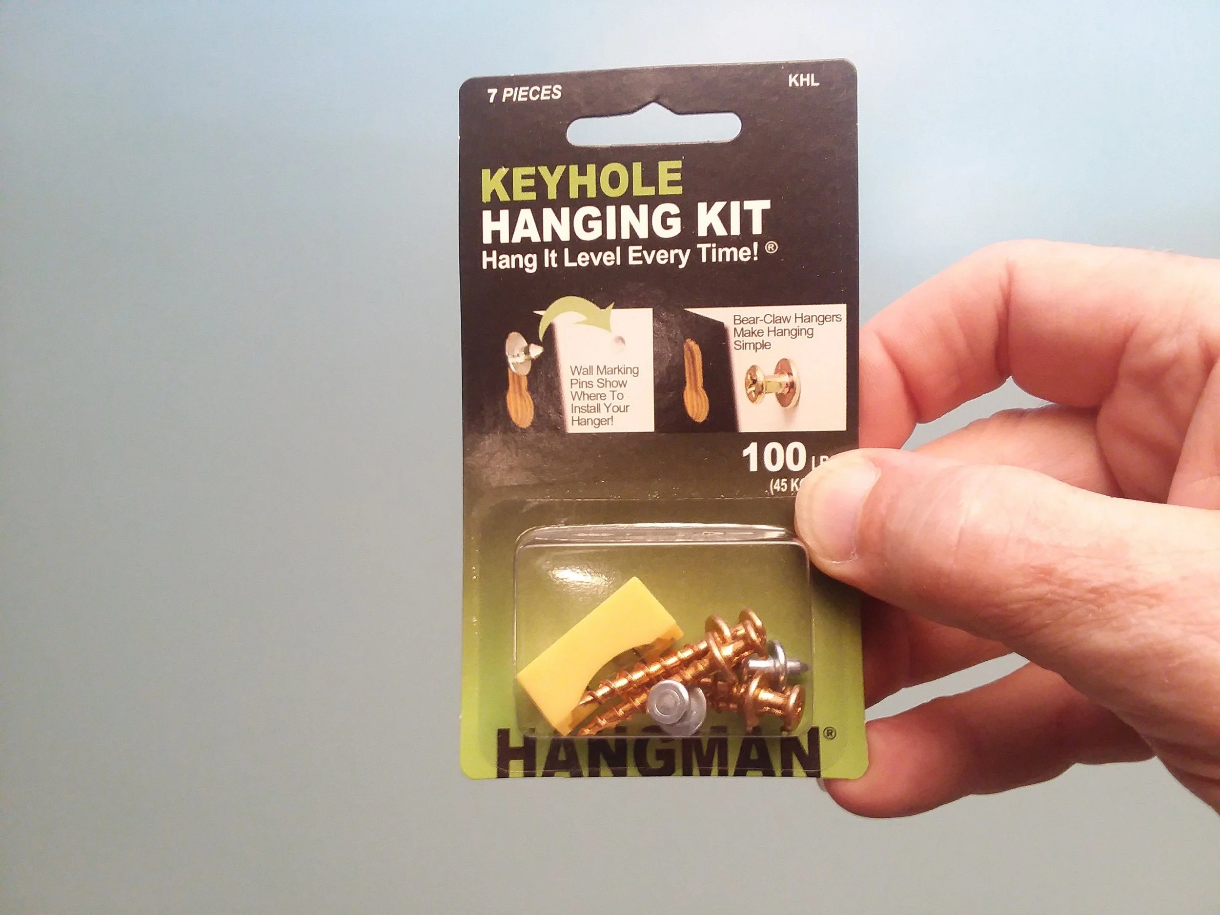Close-up of a hand holding a Keyhole Hanging Kit package with seven pieces, featuring illustrations of wall marking pins.
