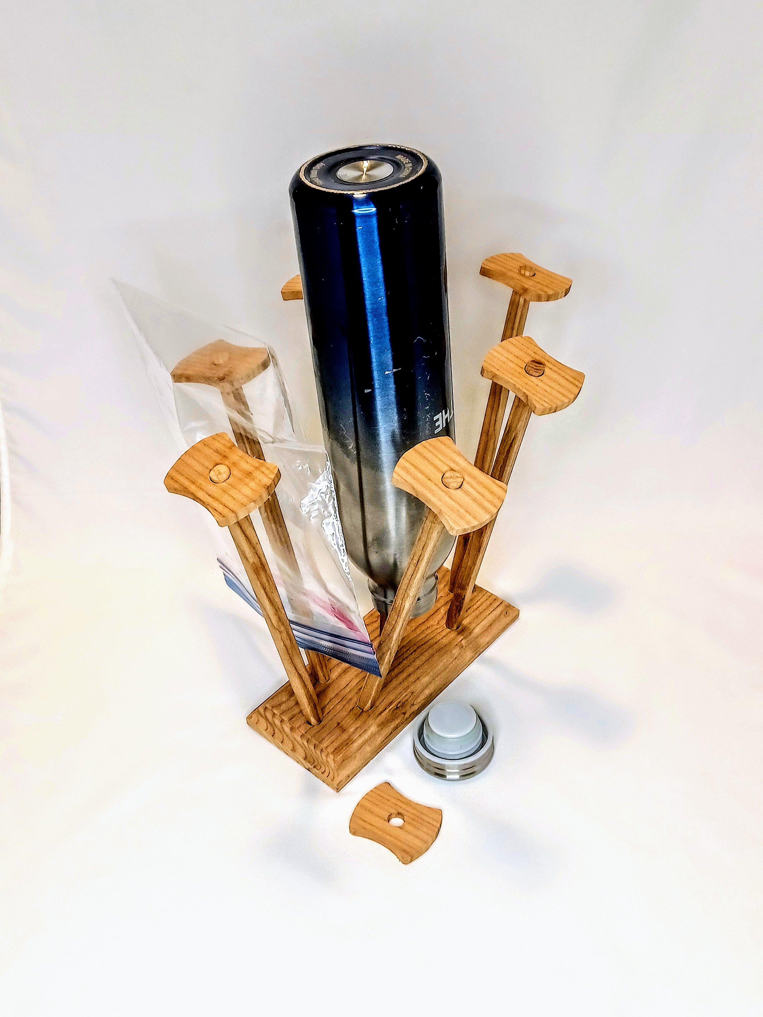 An inverted water bottle drying on a wooden stand with posts holding plastic bags and a white background.
