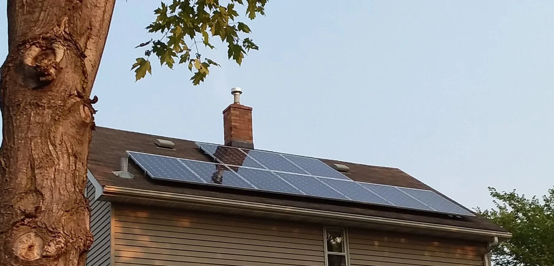 A house with a solar panel system installed on its roof and a brick chimney. A tree with green leaves is in the foreground.