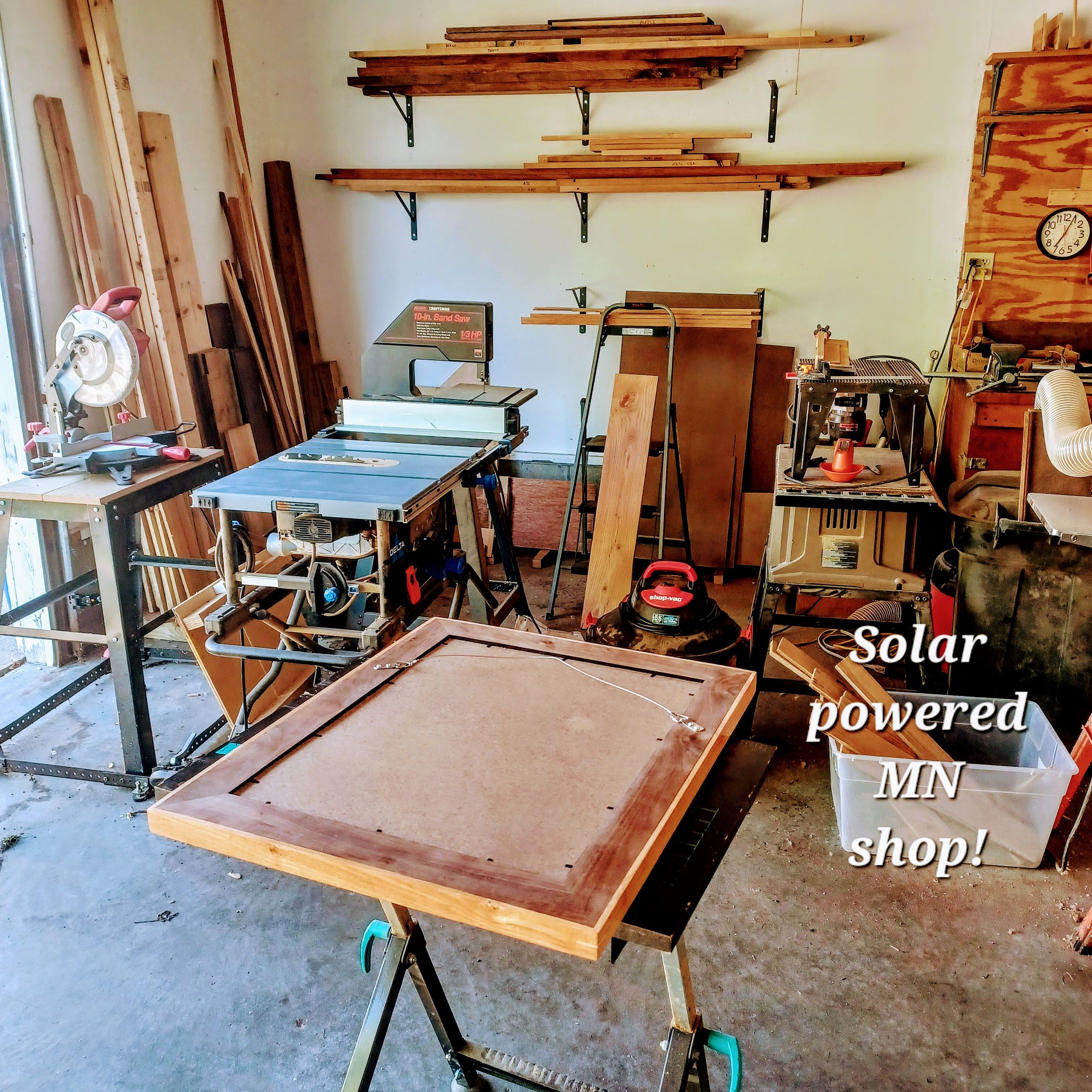 A workshop with various tools and woodworking materials. There is a table saw, several pieces of wood, shelving with more wood, and a wall clock. The space appears to be used for carpentry or woodworking projects.
