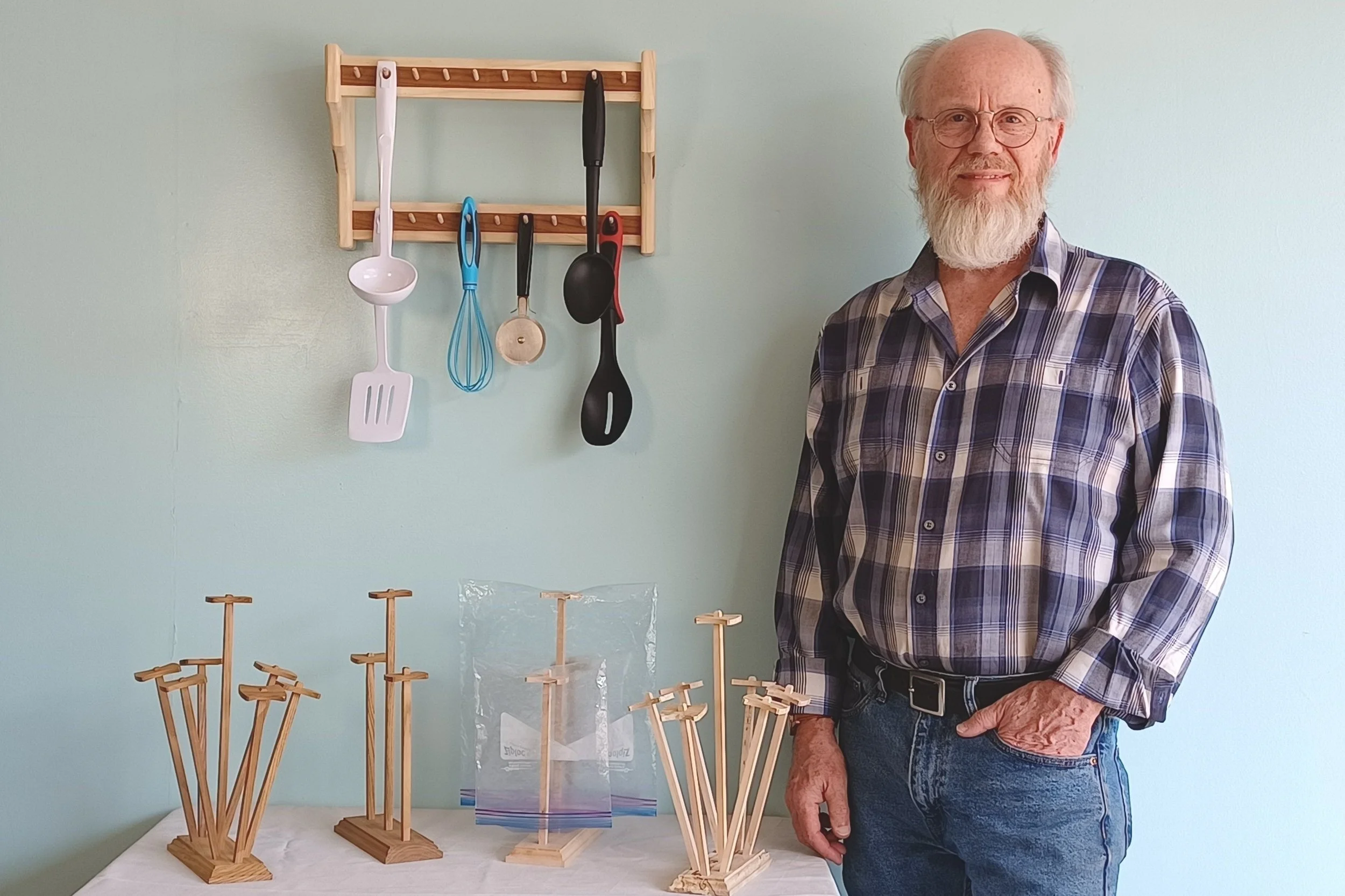 An elderly man with glasses and a beard wearing a plaid shirt standing next to a display of wooden objects on a table and a wall-mounted hook rack with kitchen utensils hanging. The background is a pale green wall.