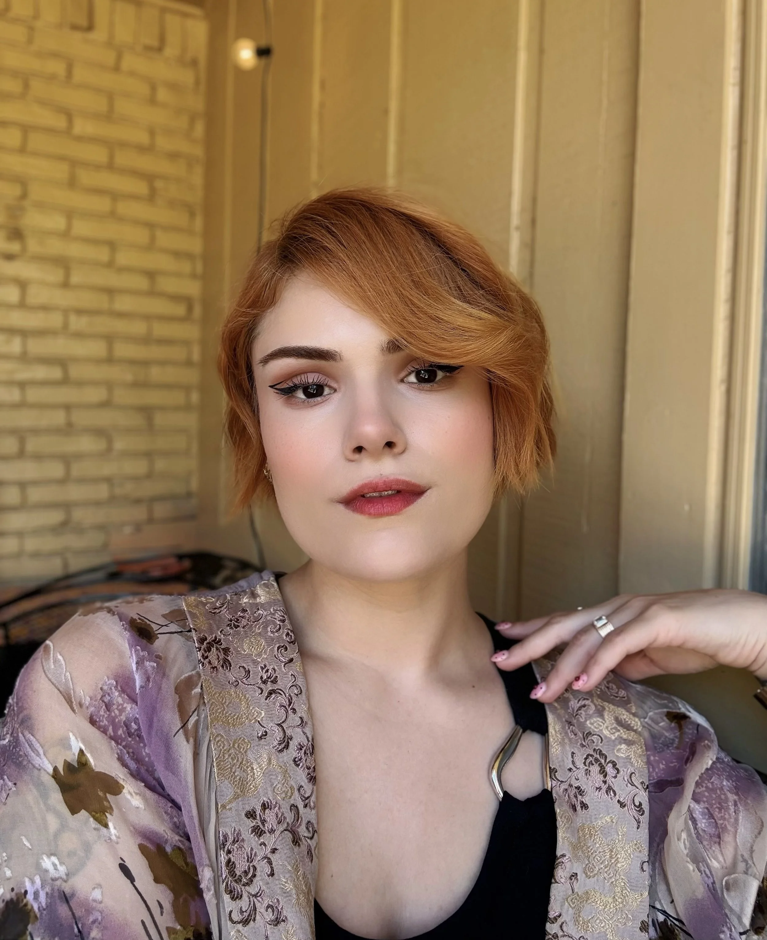Young woman with short, reddish-orange hair, wearing a white tie-dye top, layered necklaces, earrings, and has colorful tattoos on her arms, standing against a plain white background.