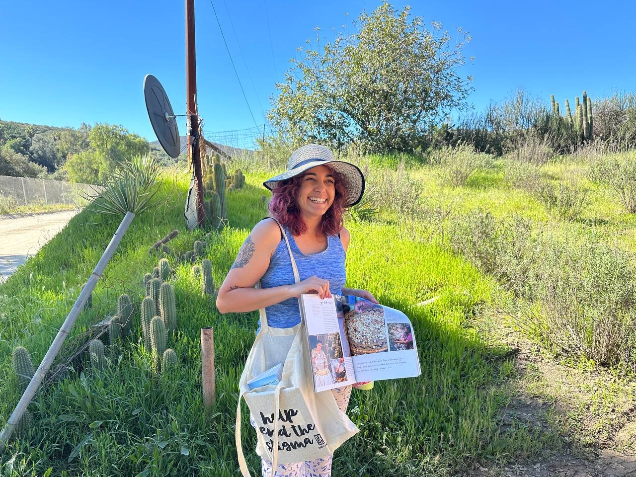 A woman wearing a sunhat and blue tank top standing in a grassy area with cacti and bushes, smiling and holding an open magazine.