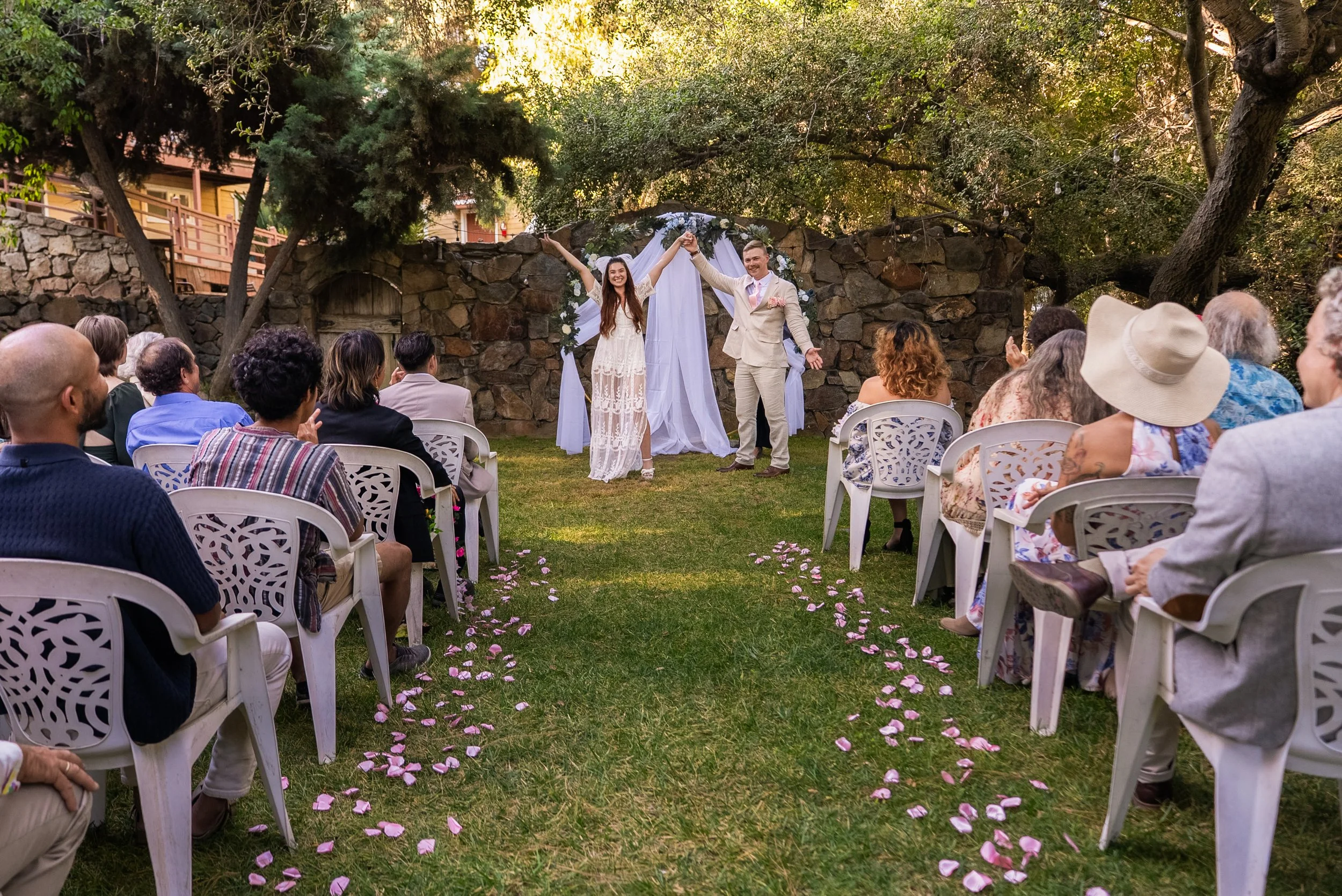 A couple and their wedding guests celebrating outdoors under trees, with the couple holding hands up in the air near a decorated backdrop, and pink flower petals scattered on the grass aisle. DIY wedding venue in San Diego.