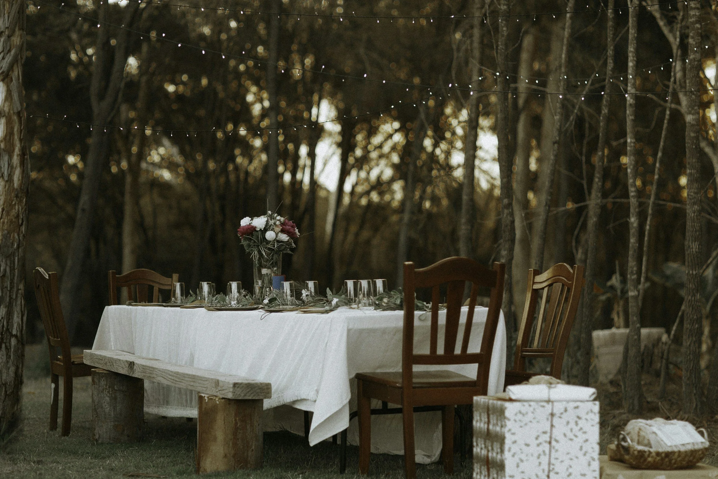 A table set for a meal outdoors in a wooded area with string lights overhead. The table has a white tablecloth, a floral centerpiece, wine glasses, and place settings. Wooden chairs surround the table.