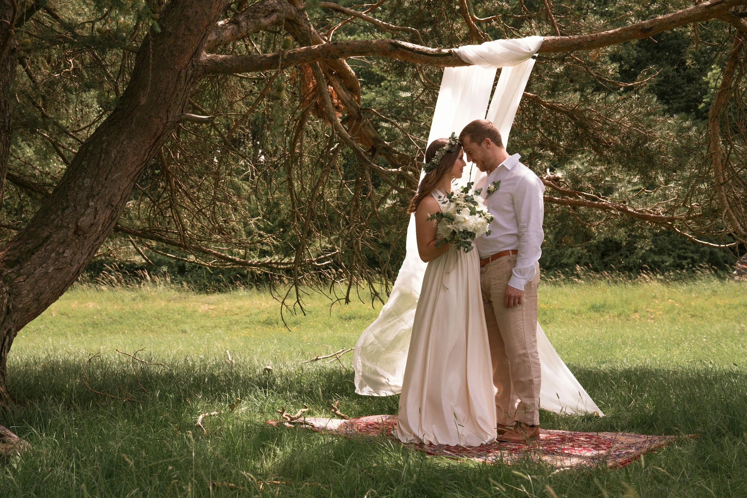 A couple dressed in wedding attire standing closely under a tree during their outdoor wedding ceremony, with a white fabric backdrop hanging from the branches and a patterned rug on the grass.