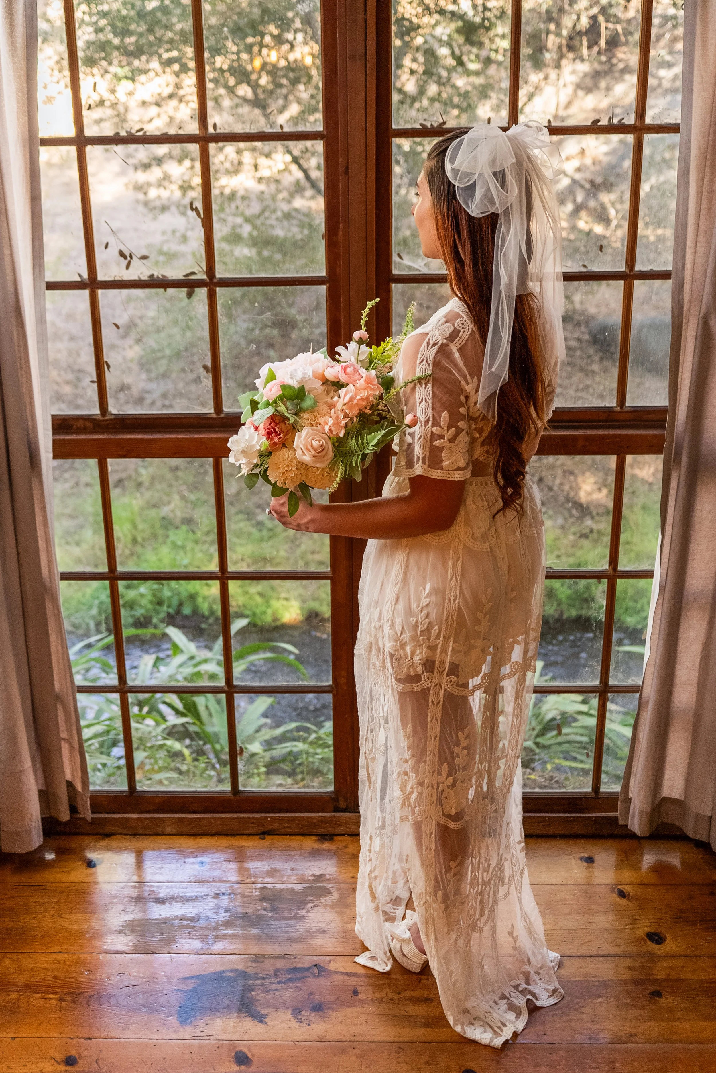 A woman in a wedding dress holding a bouquet of flowers, standing by a wooden and glass door, with sunlight filtering through trees outside.