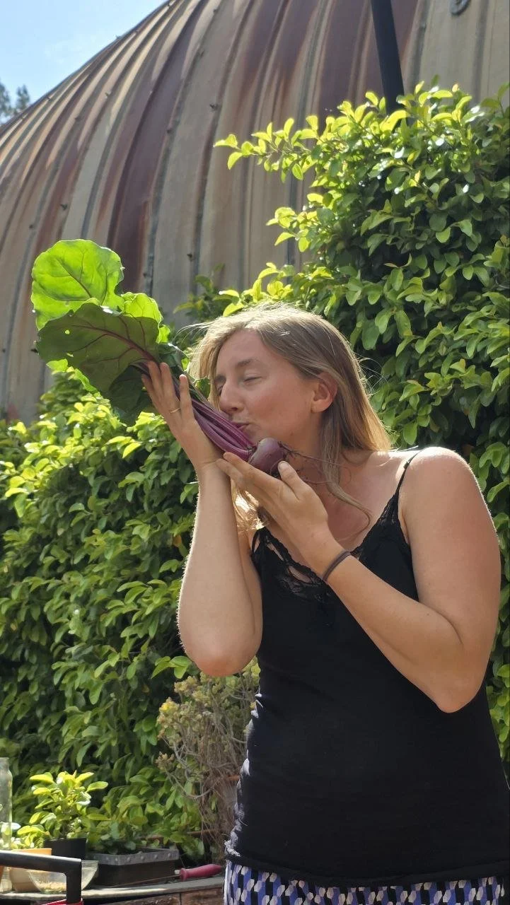 A woman in a black tank top and patterned skirt kisses a large purple and green vegetable, likely a beet, outdoors with green foliage and a rust-colored shed in the background. Backyard garden in a sustainable community.