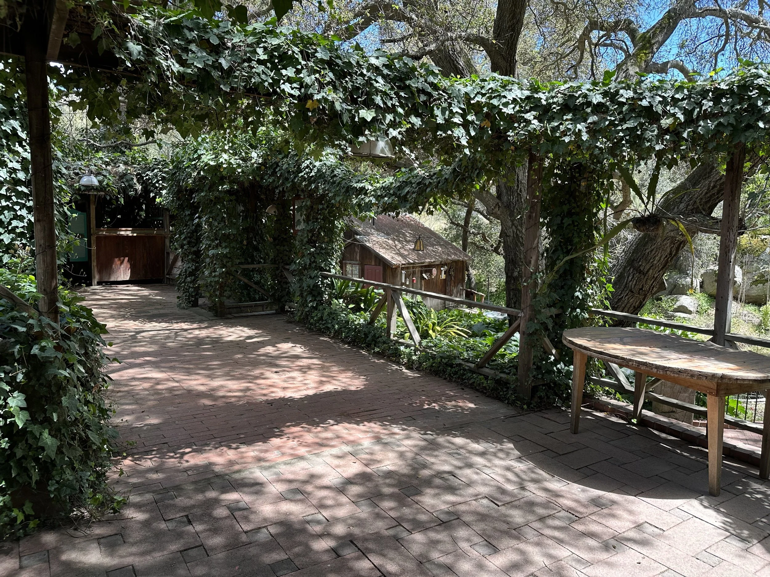 A shaded outdoor patio area with brick pavers, surrounded by lush green foliage and trees, with small wooden structures and a railing.