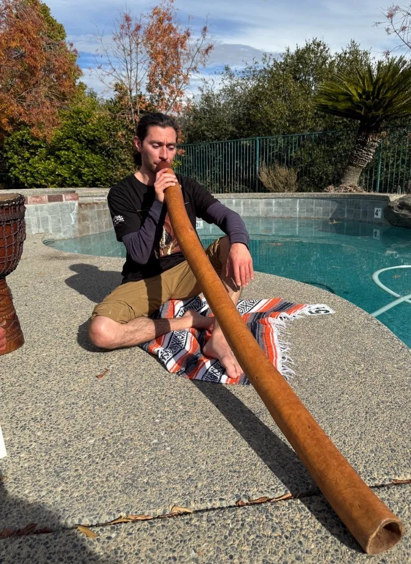 A man sitting cross-legged on a colorful striped towel by a swimming pool, playing a didgeridoo, with trees and sky in the background. A resident in a sustainable community.