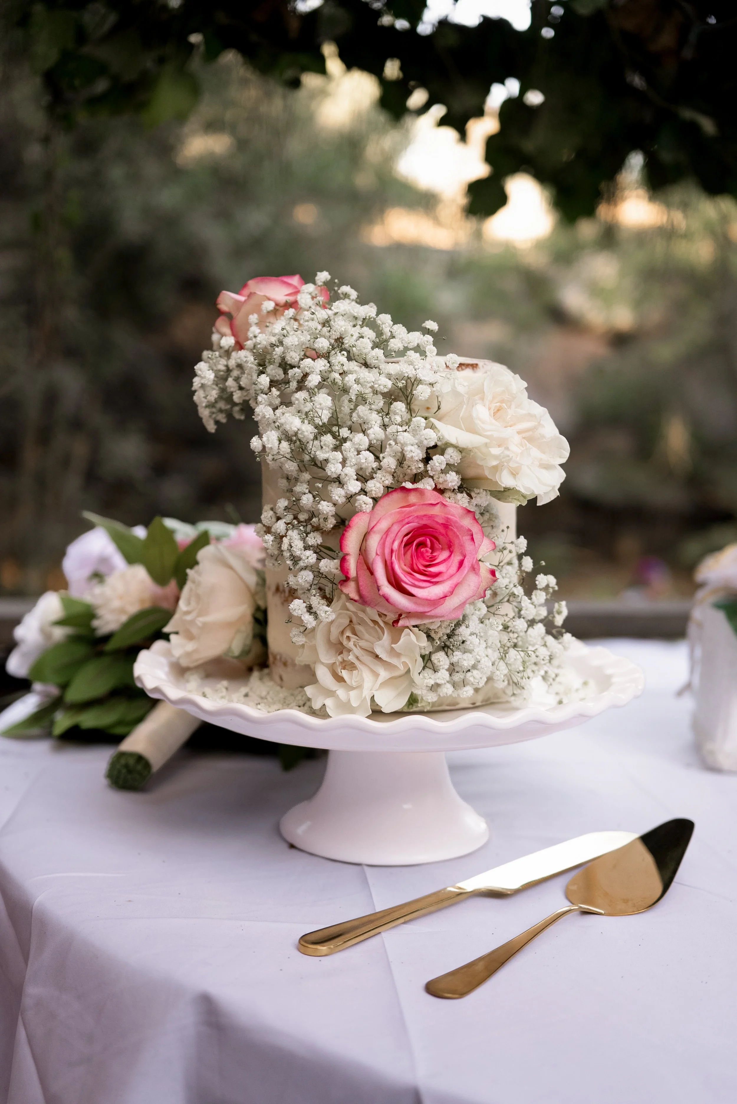 Wedding cake with pink and white roses and baby's breath flowers on a white cake stand, set on a table with gold utensils and a white tablecloth, outdoors at sunset.