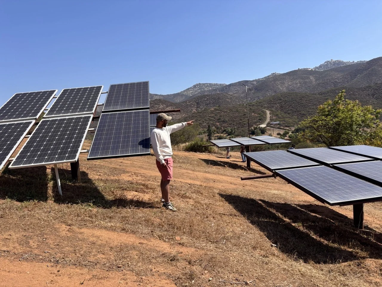 A person standing in front of solar panel arrays on a hillside, pointing towards the mountains in the background under a clear blue sky.