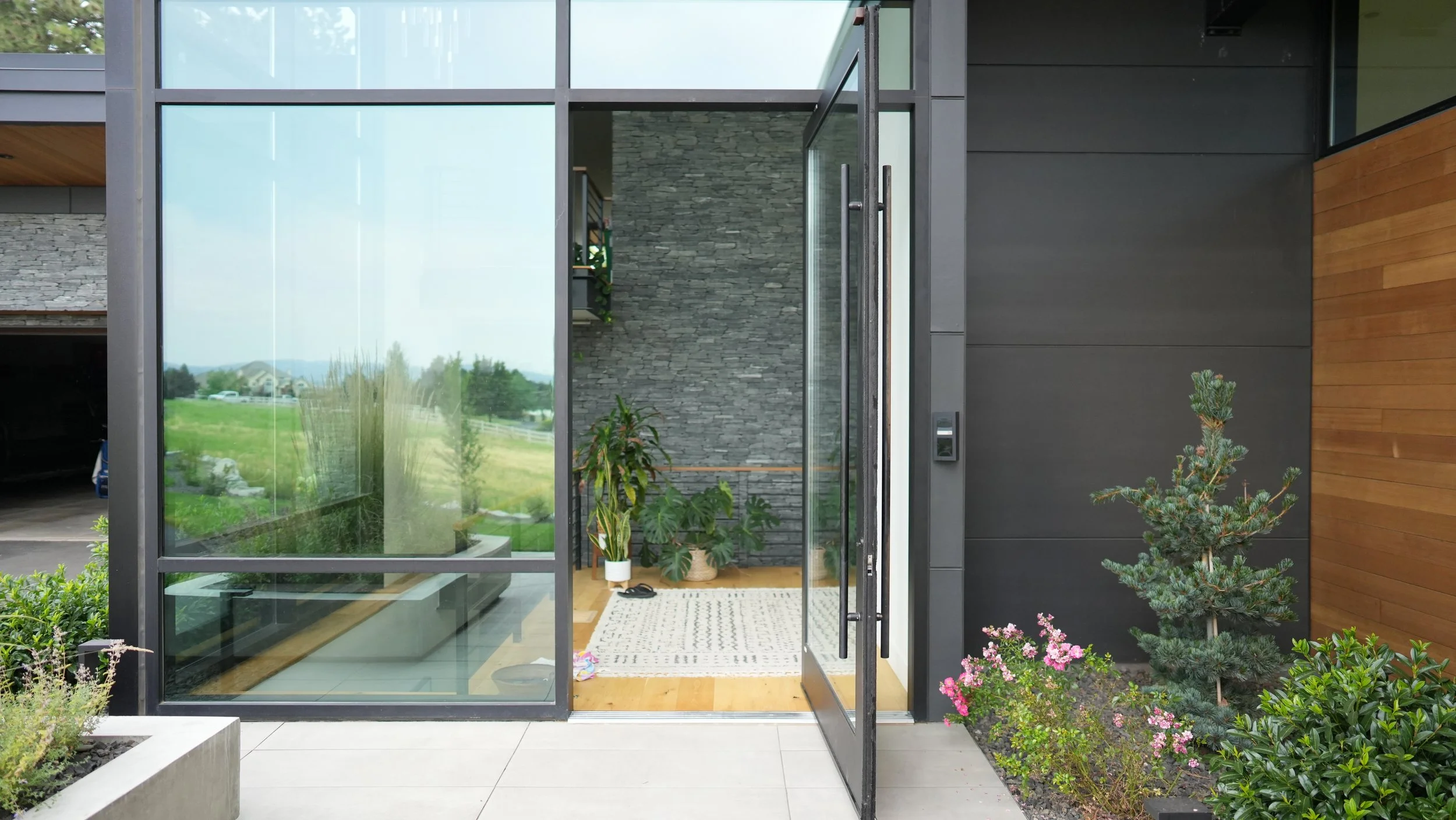 Entrance to a modern house with glass sliding door, indoor plants, and outdoor garden with bushes and a small pine tree.