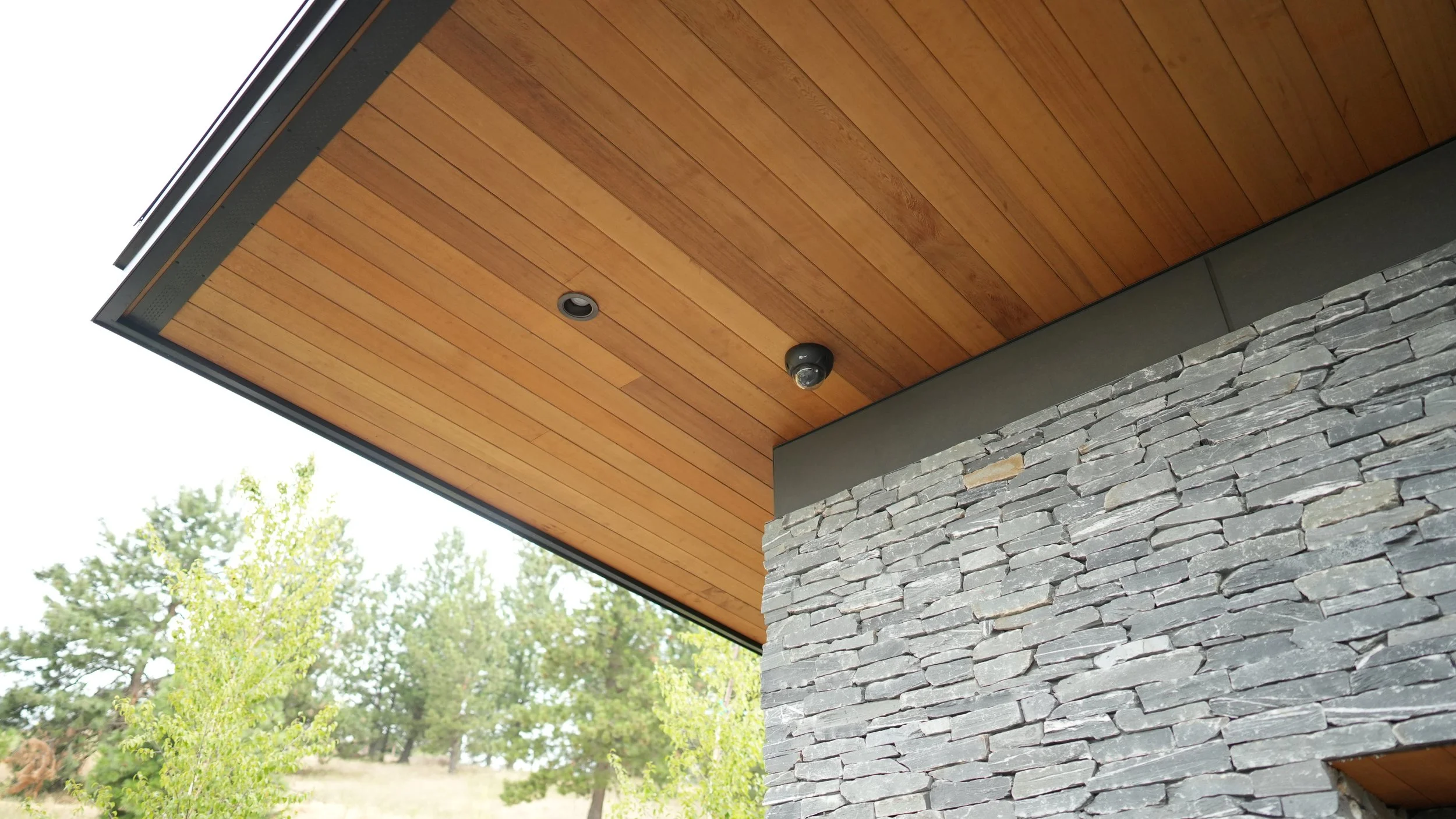 Close-up of modern house exterior with wooden ceiling, gray stone wall, and outdoor security camera, with trees in the background.