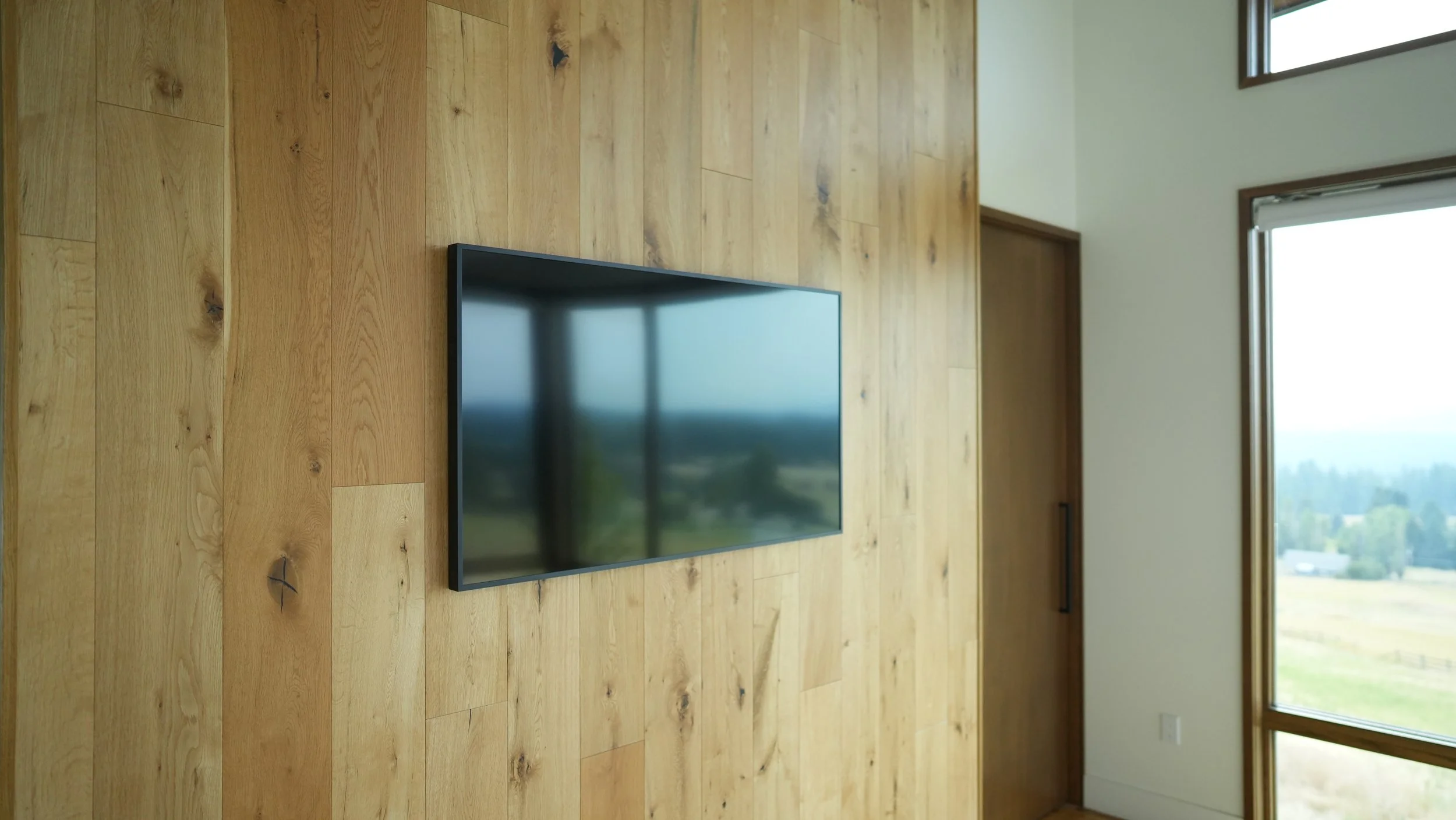 Flat-screen TV mounted on a wooden wall in a modern room with large windows showing an outdoor landscape.