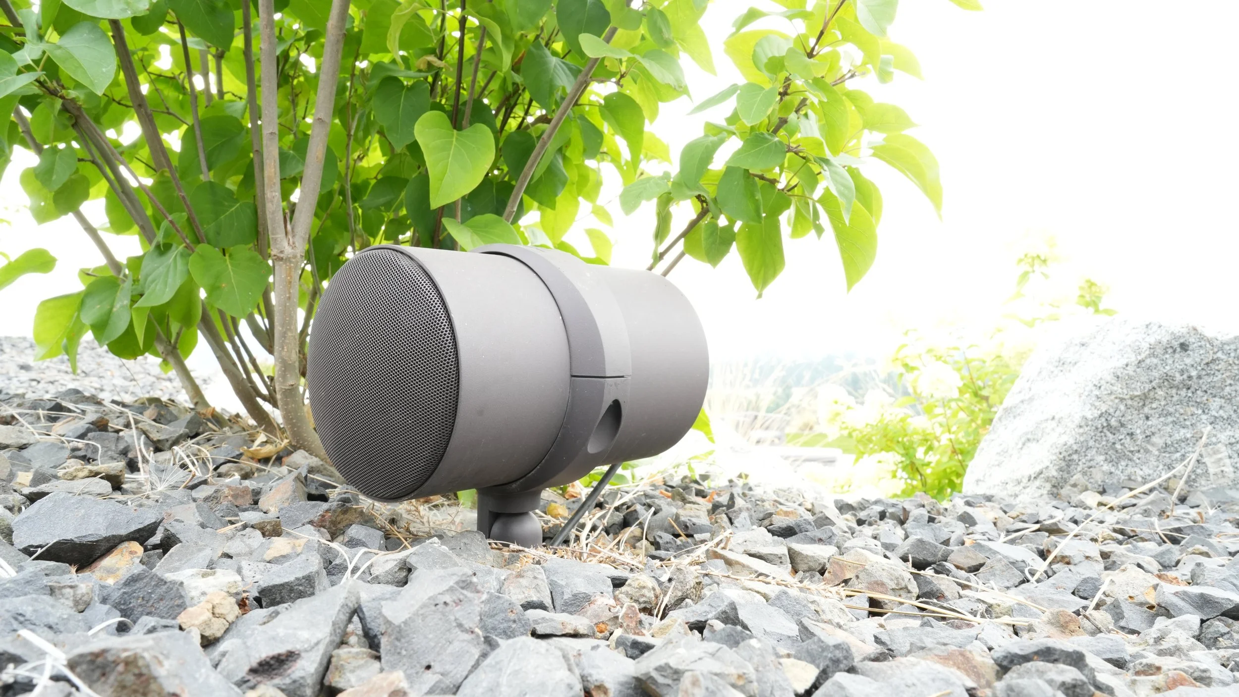 Wireless outdoor speaker placed on gravel next to green leafy bushes and rocks in sunlight.