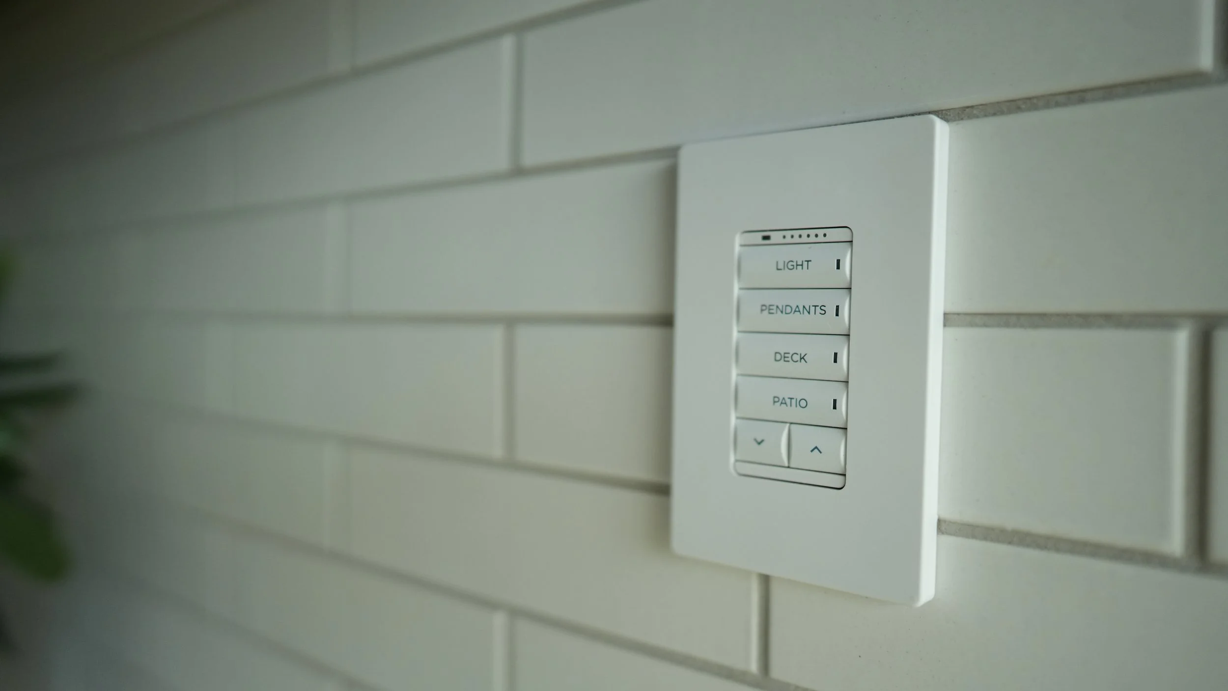Remote control panel mounted on a light-colored brick wall with buttons labeled for controlling lights, pendants, deck, and patio.