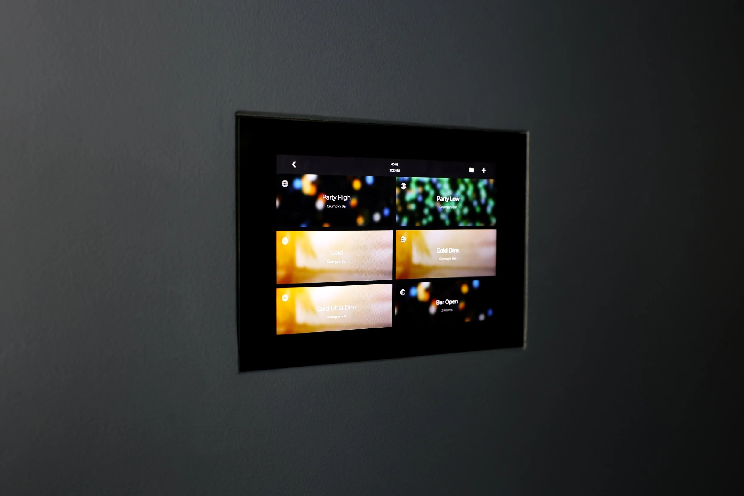 A digital touch screen control panel embedded in a gray wall, displaying scene options for lighting or entertainment, including 'Party High,' 'Party Low,' 'Gold,' 'Gold Dim,' 'Gold Ultra Dim,' and 'Bar Open'.
