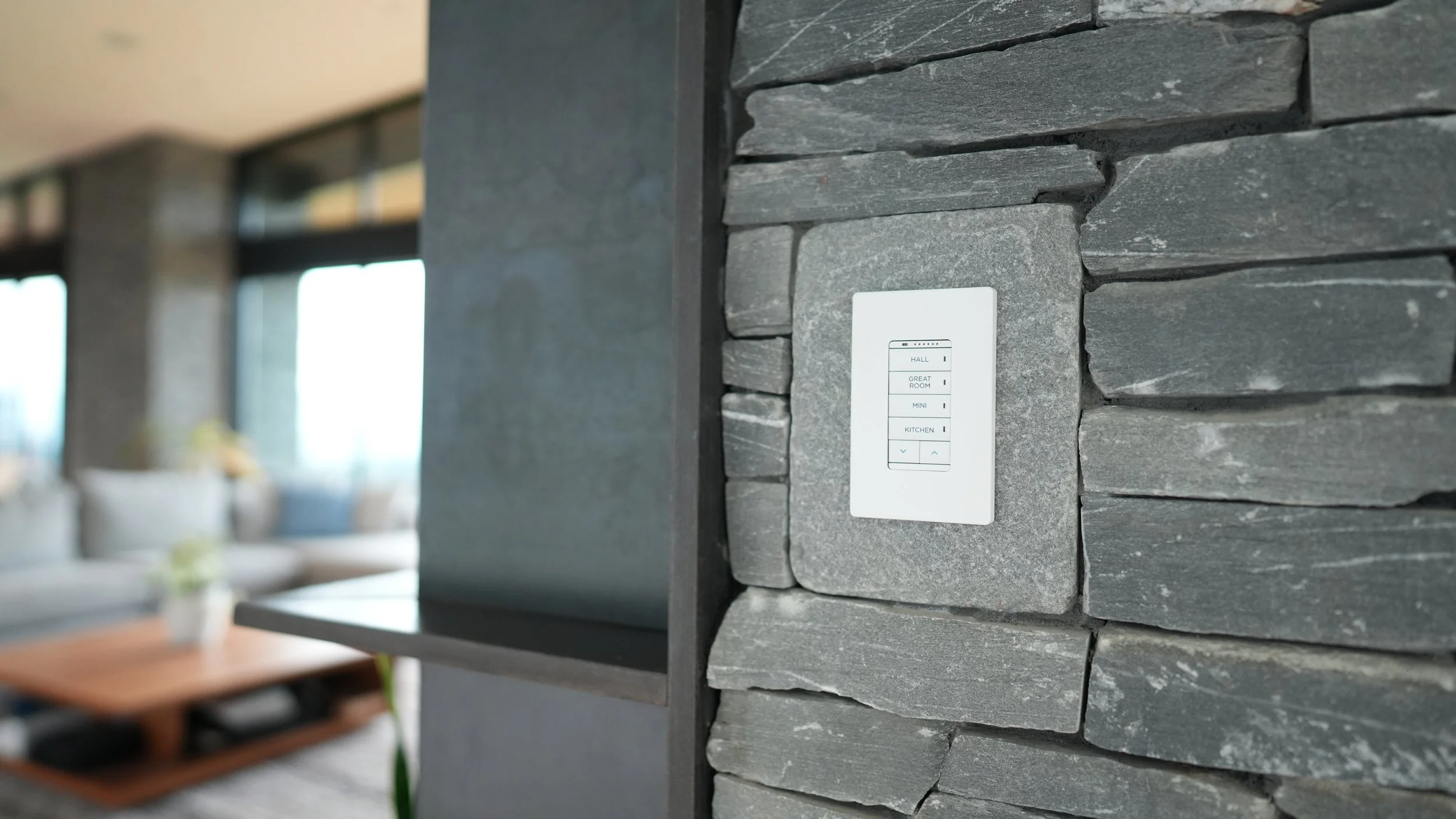 A modern wall-mounted control panel with a white faceplate and black buttons, installed on a textured dark gray stone wall in a contemporary living space.