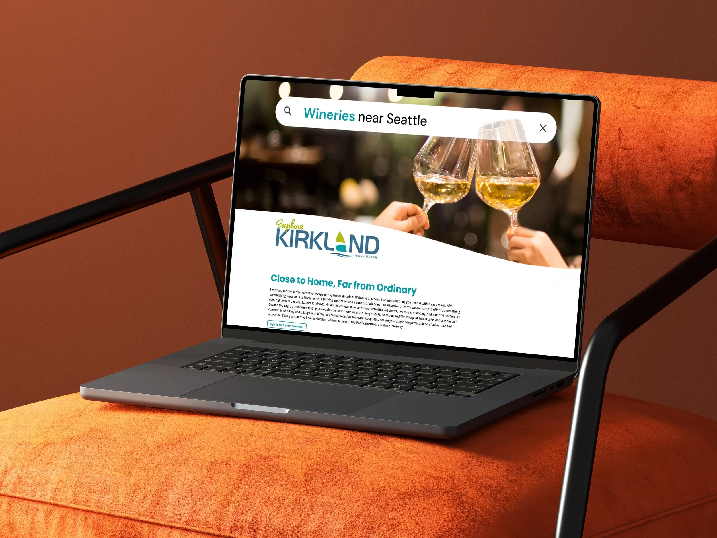 Laptop on an orange cushion displaying a website for Kirkland with an image of two people clinking glasses of white wine.