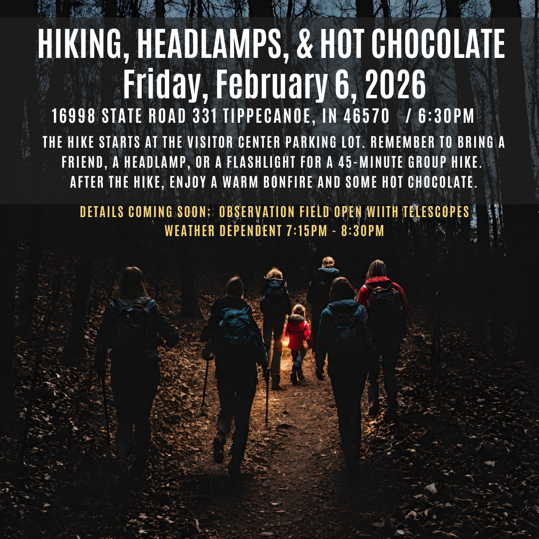 A group of hikers walking on a trail in the woods during dusk, wearing backpacks and outdoor clothing, with tall trees around them.