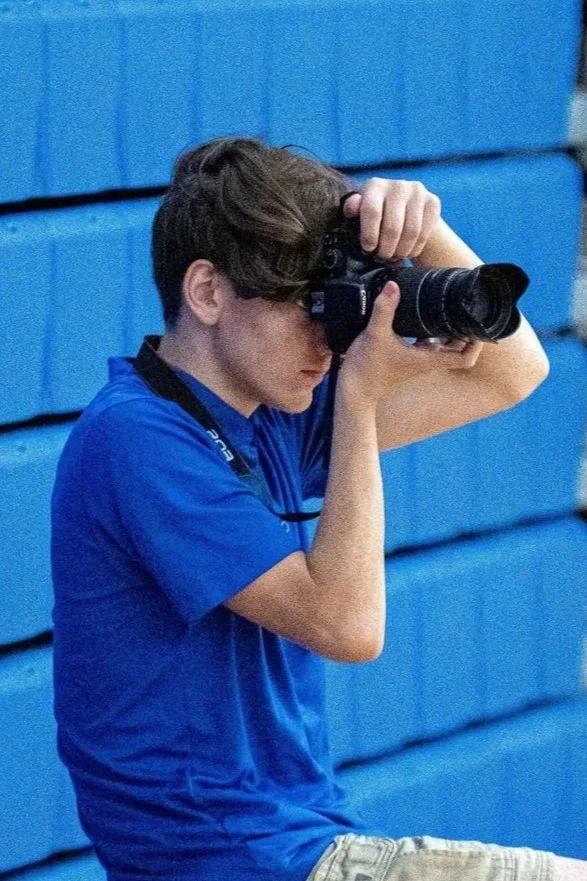 A young man with brown hair in a blue polo taking a photo with a camera, sitting against a blue bleachers.