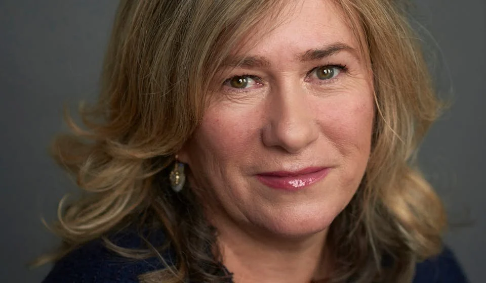 A close-up photograph of author and journalist Sonia Purnell, who is looking at the camera and smiling with a closed mouth. Sonia has caramel blonde hair and hazel eyes, and is wearing gold earrings and a black top.