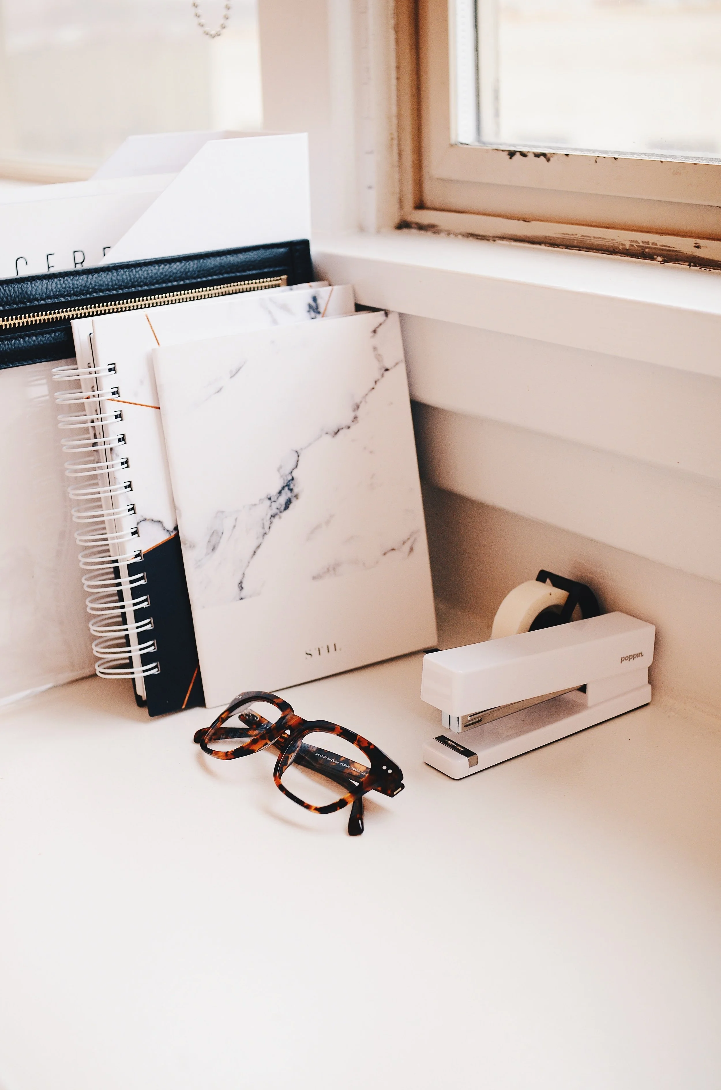 Desk with notebooks, glasses, and a stapler near a window.